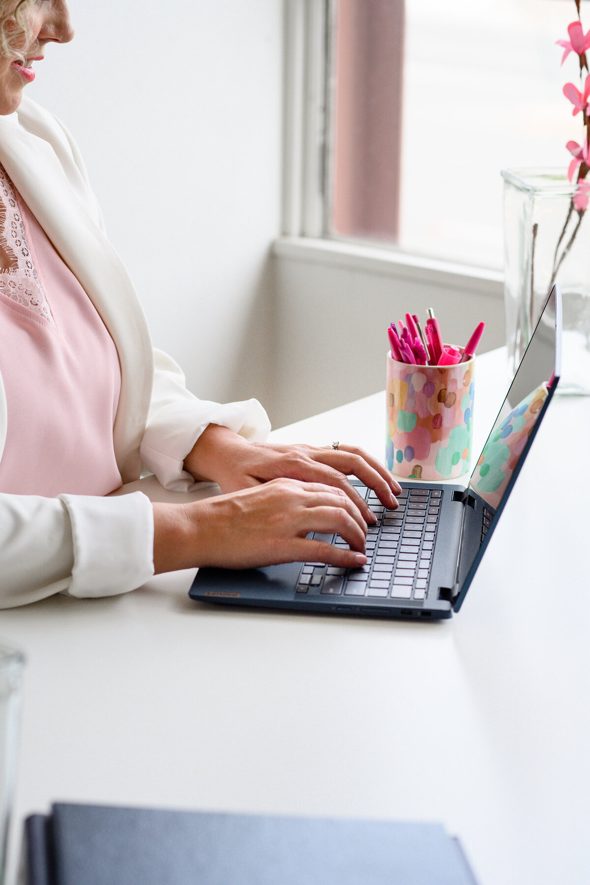 photography branding for a woman in a pink shirt and blazer typing on her black laptop at her white desk with pens in the background