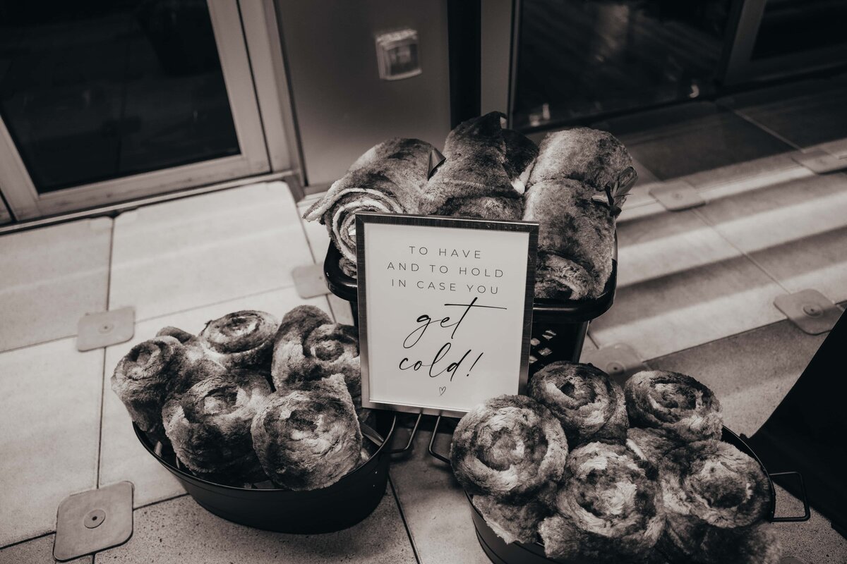 Black and white photo of baskets containing rolled-up blankets with a sign reading "To have and to hold in case you get cold!" at an Iowa wedding.