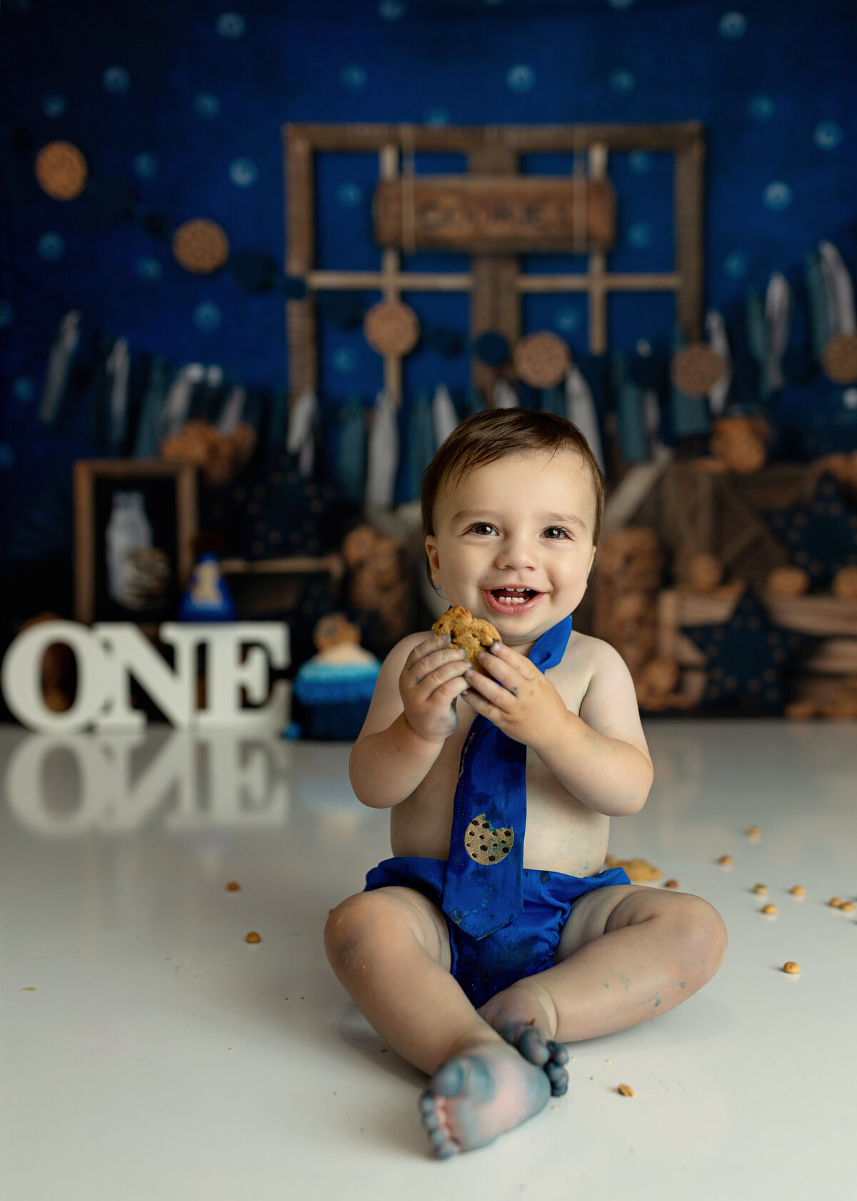 A toddler boy in a blue tie with a cookie sits in a studio eating a cookie with a smile
