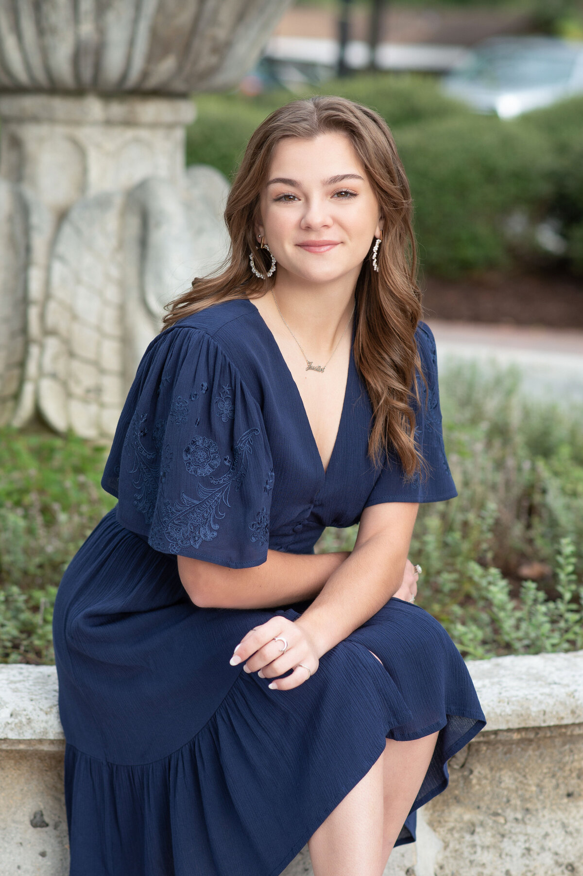 High school senior girl in a blue dress sits on ledge of fountain.