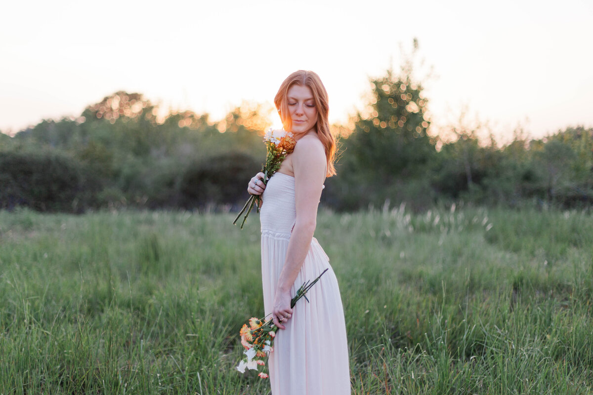 Young woman holds flowers in both hands in a field with tall green grass while the sun sets
