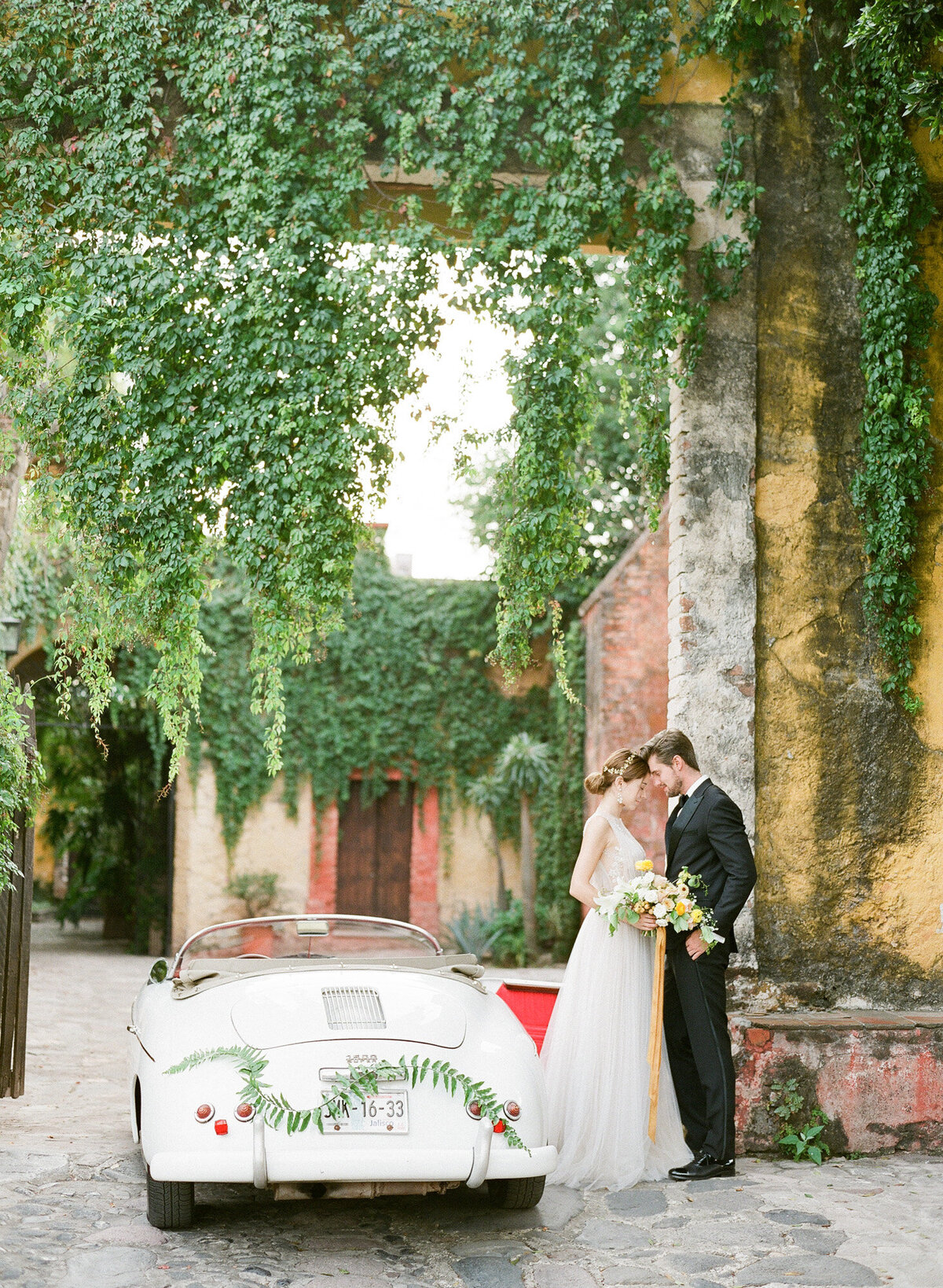 Elegantly dressed bride and groom lean towards each other beside a vintage wedding car in front of walls engulfed by leaves.
