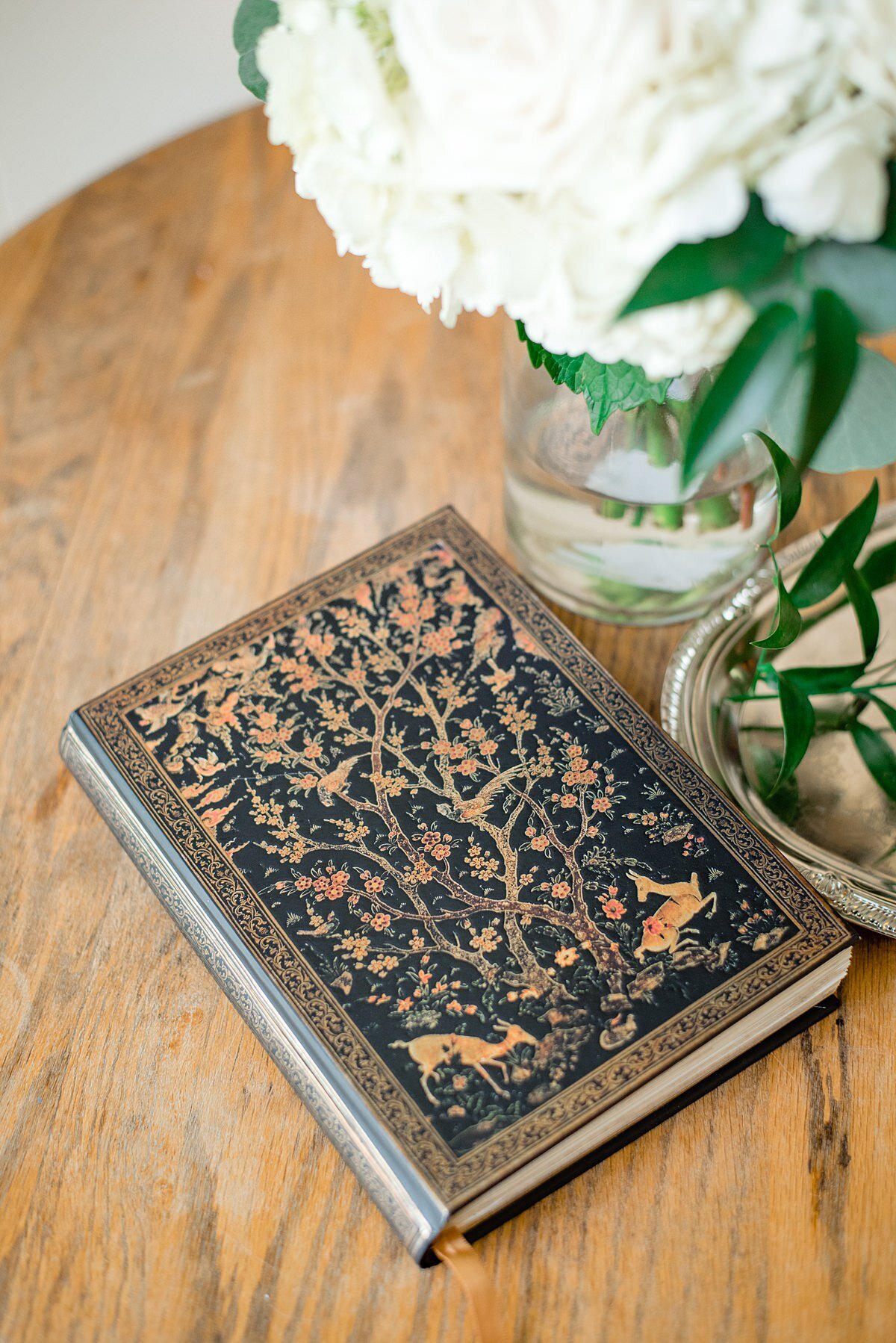 The officiant's ceremony book sits on a wooden table. The cover of the book is embossed with a gold leaf tree of life and two deer frolicking beneath the tree. To the right of the book is a silver tray and a glass vase with the bride's white bouquet.