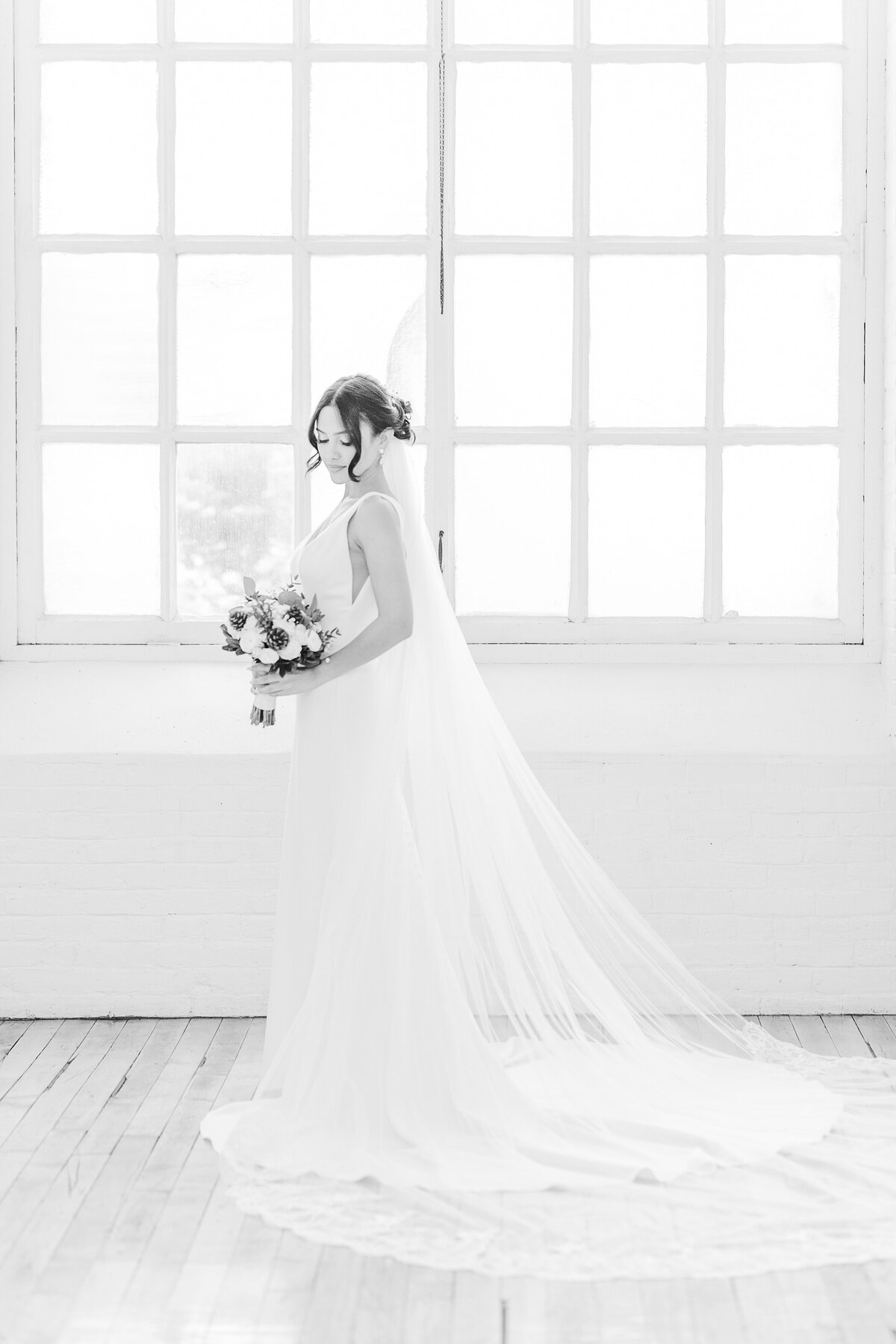Black and white image. A bride stands in front of large factory windows at Boylston Rooms in Easthampton, MA. The bride is standing side profile to the camera with her bouquet in front of her. She is looking over her shoulder and head town. Captured by MA wedding photographer Lia Rose Weddings