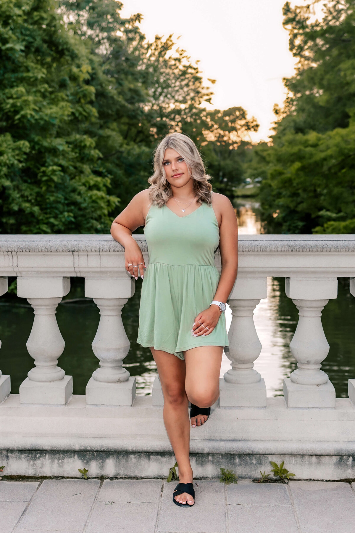 A blonde haired high school senior girl leaned against a concrete railing with trees and a pond in the background at sunset