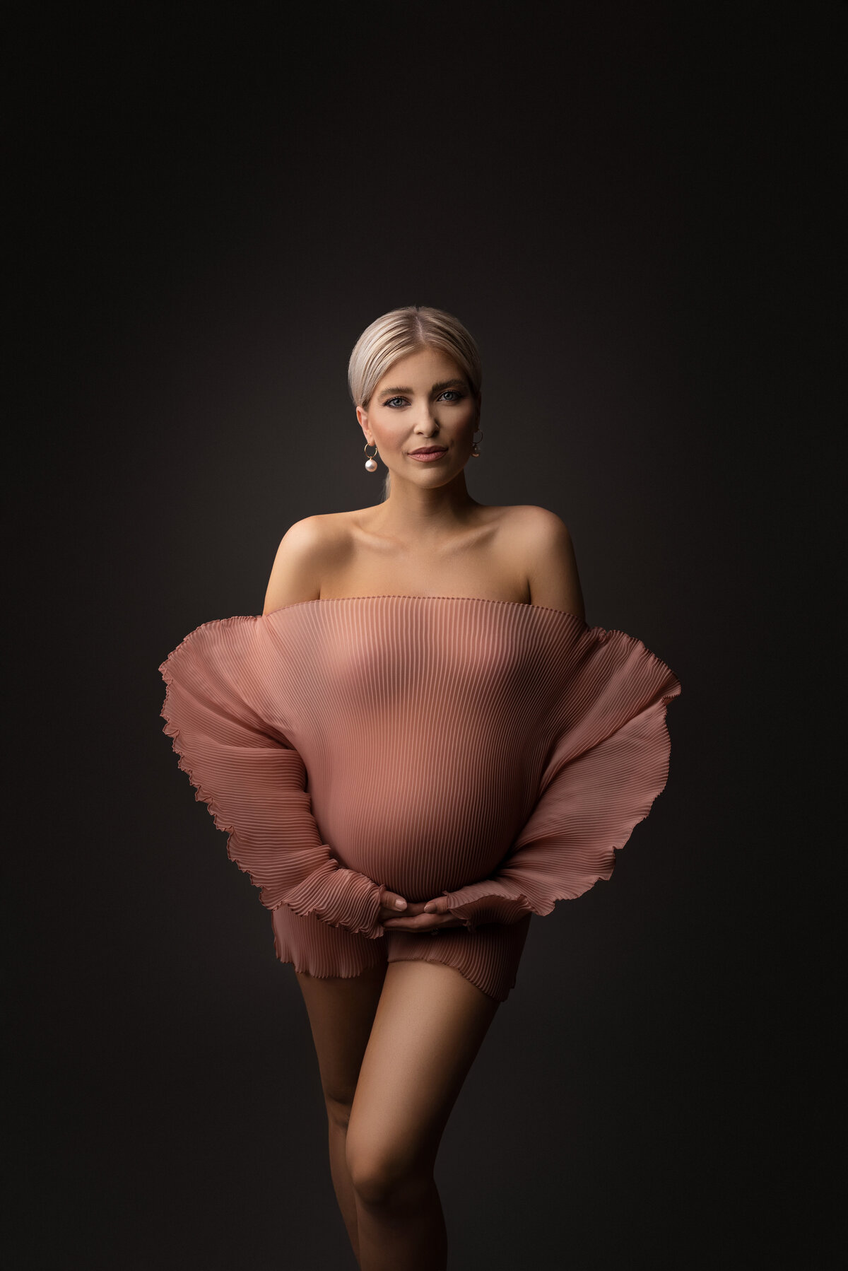 Woman poses for fine art maternity photos with New Jersey's best maternity photographer Katie Marshall. Woman is standing facing the camera in an off-the-shoulder dusty rose organza photoshoot mini dress with long, embellished sleeves. Her front knee is bent in front of her and both ands are resting gently underneath her baby bump.  The woman's hair is tied back in a low bun and has a closed mouth smile. The light is focused on the left half of her body, creating shadows and depth to her right.