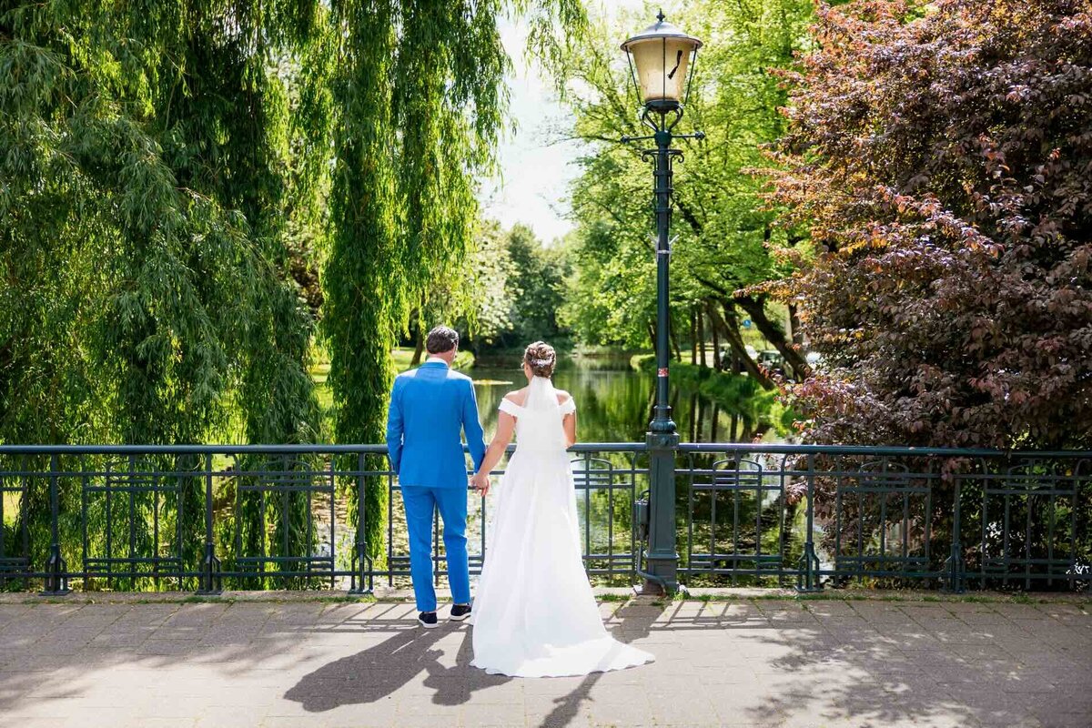 Bruid en bruidegom lopen hand in hand met rug naar camera, kijkend over de gracht tijdens hun bruiloft