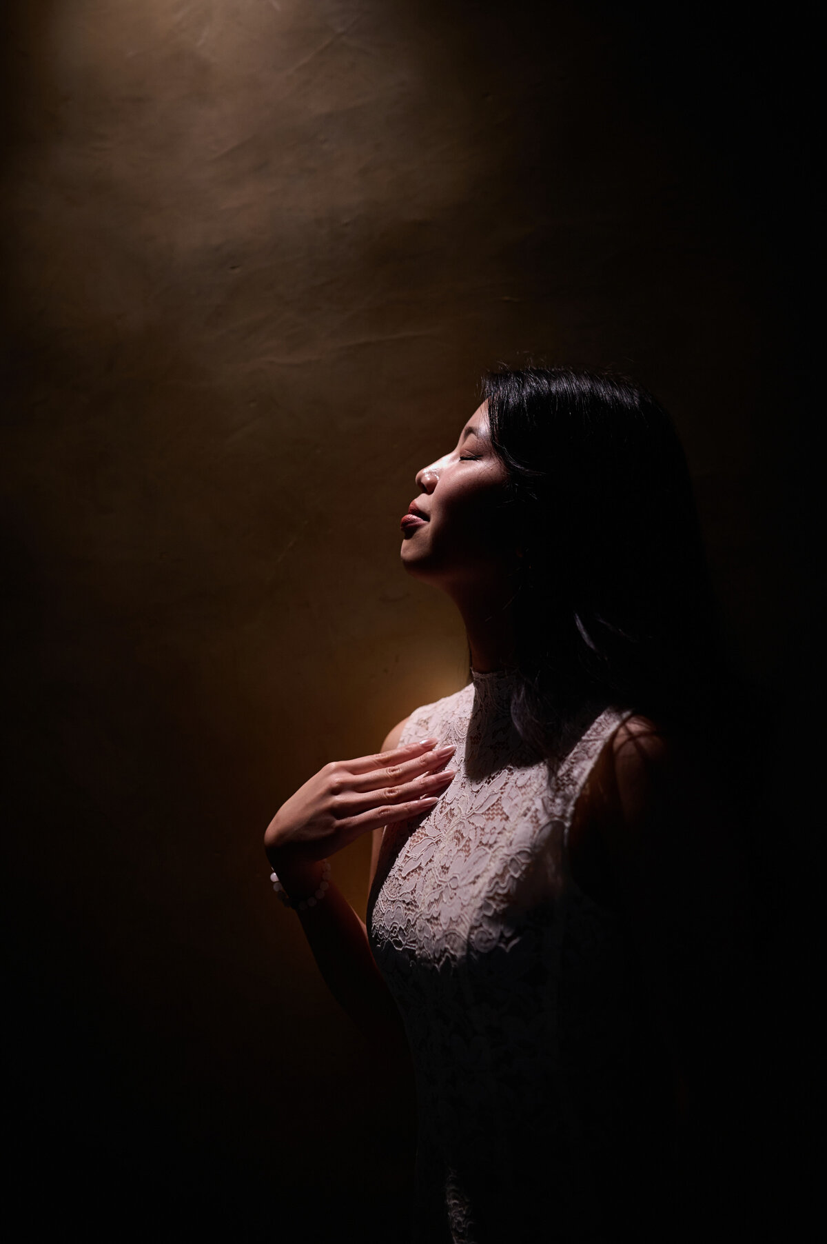 Vietnamese bride is standing in soft light. Her eyes are closed her hand resting gently on her chest