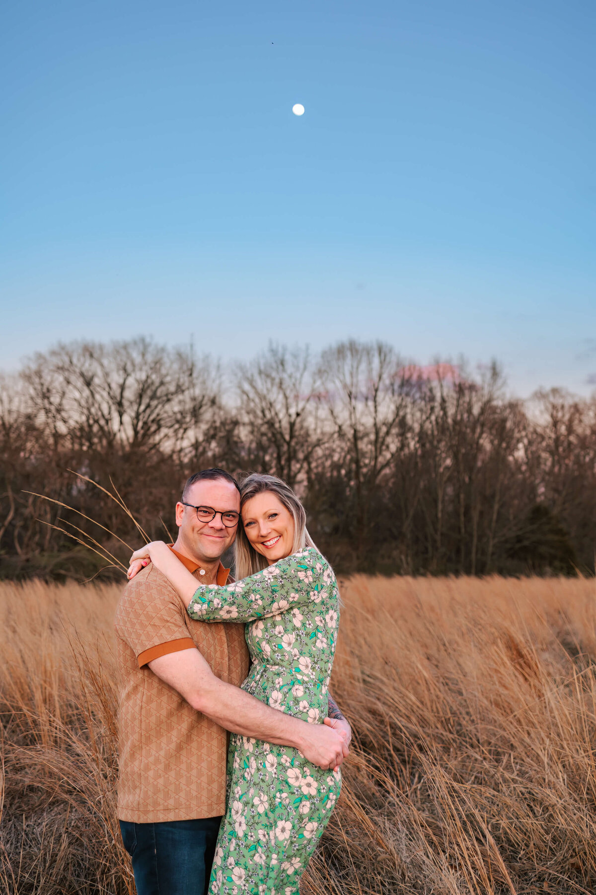 A man and woman are standing together in a field of tall grass  with their arms around each other smiling at the camera. The moon is showing in the blue sky