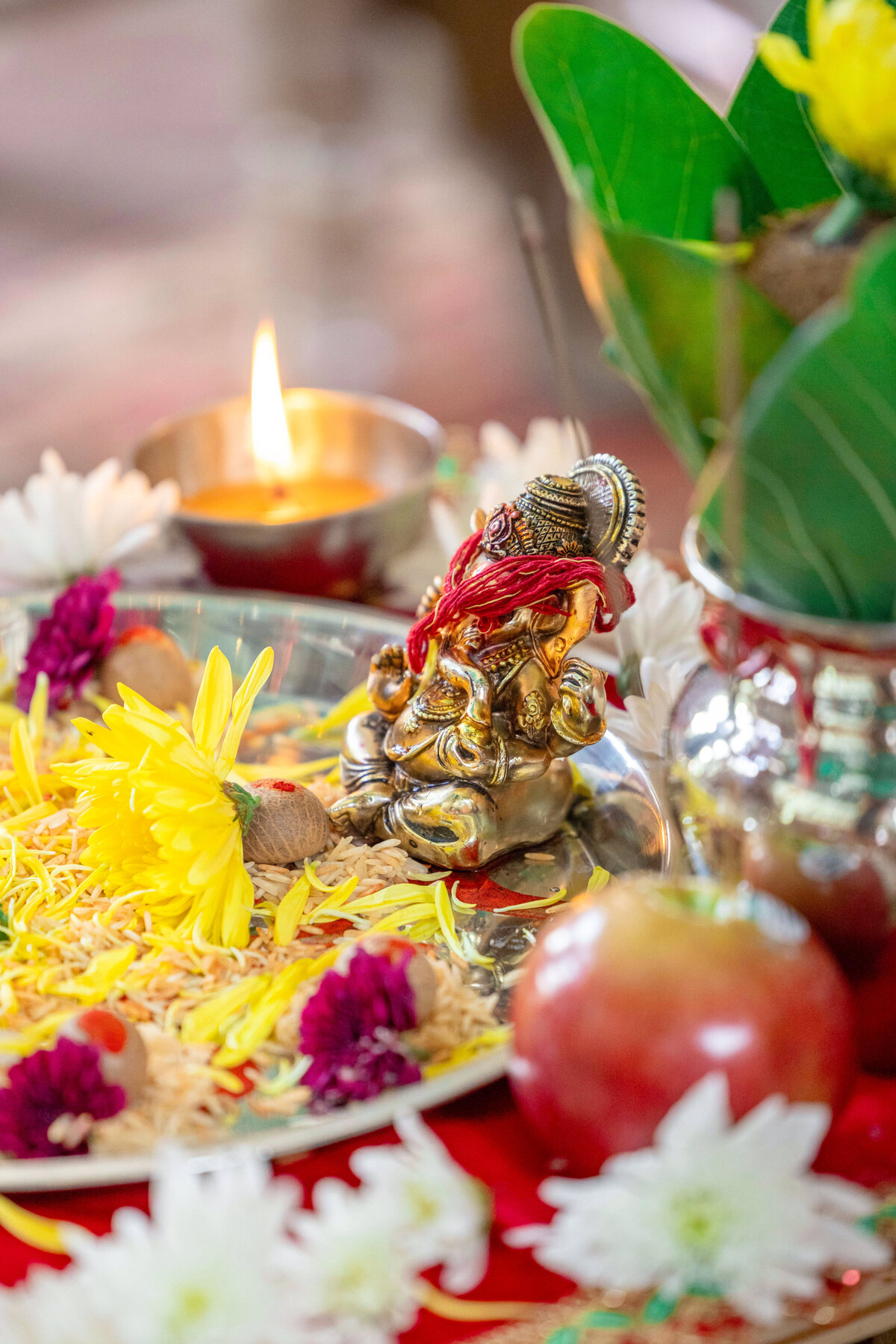 A small Ganesha idol surrounded by yellow and white flowers, a red apple, and other ritual items on a decorative tray. A lit candle sits nearby, adding a warm glow to the scene.