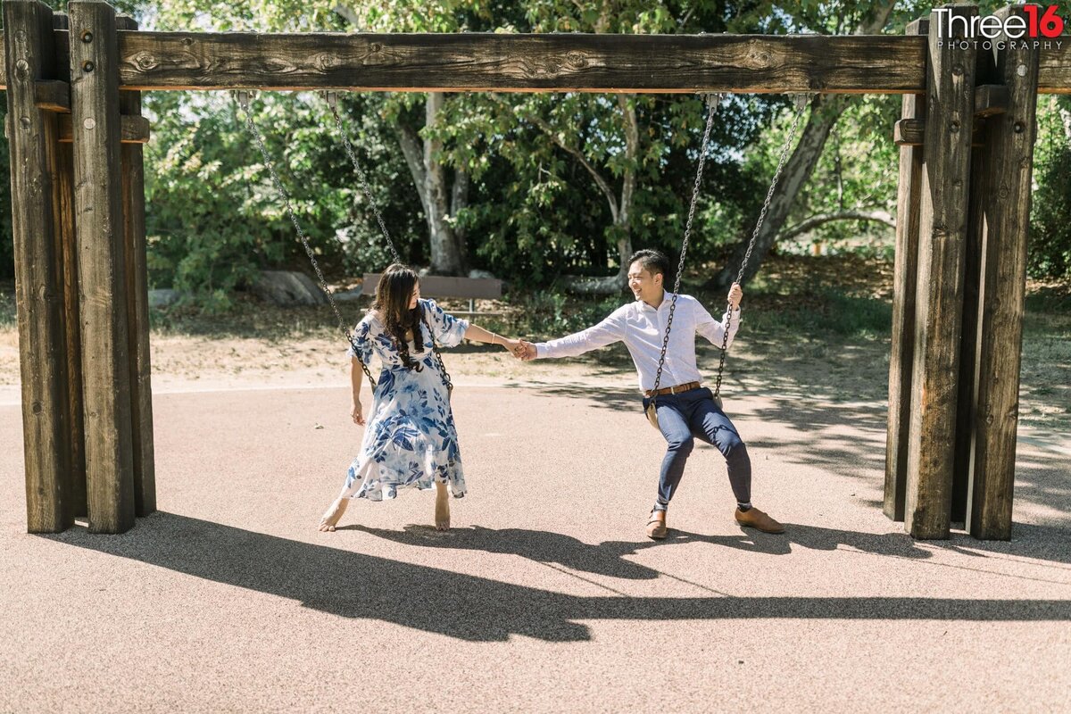 Engaged couple hold hands while swinging on a kid's swing set