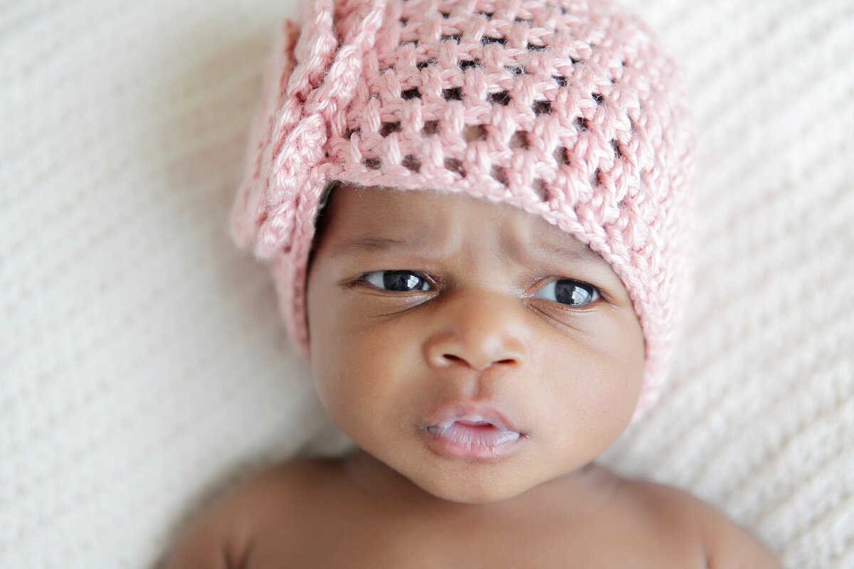 A newborn with eyes wide open in a pink crocheted hat on a white blanket.