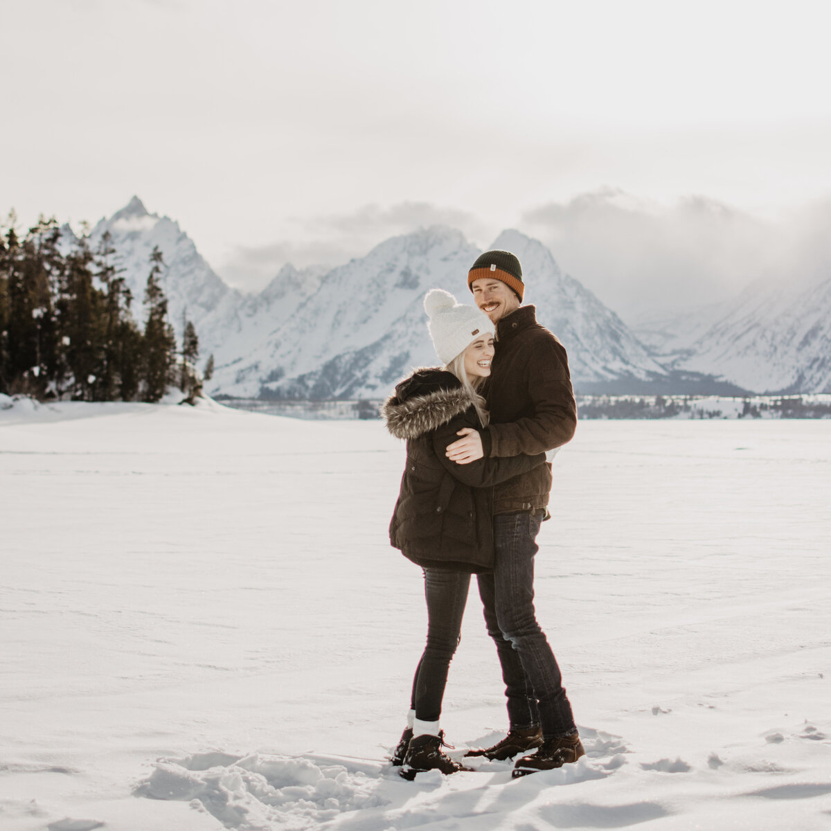 jackson hole photographers captures engaged couple hugging during their winter engagement session in Jackson Hole with the Grand Tetons behind them while it is snowing