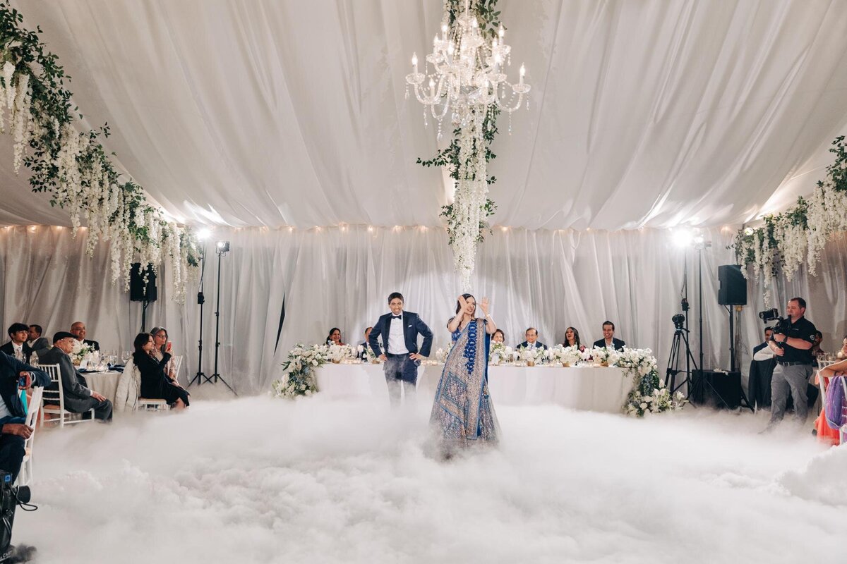 A bride and groom stand at a decorated head table surrounded by guests and a foggy effect at a wedding reception.