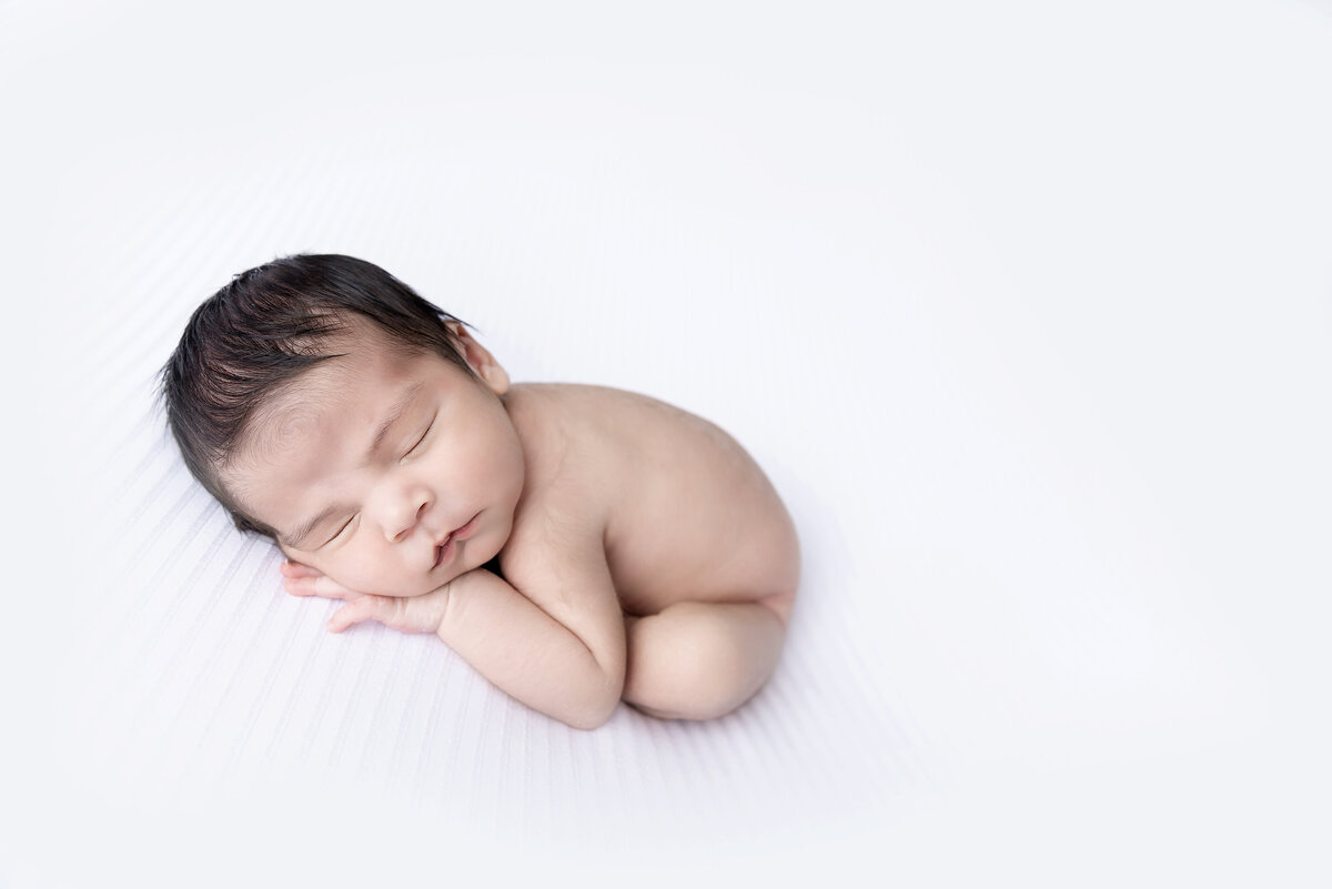 A newborn baby sleeps in froggy pose on a white blanket with dark hair