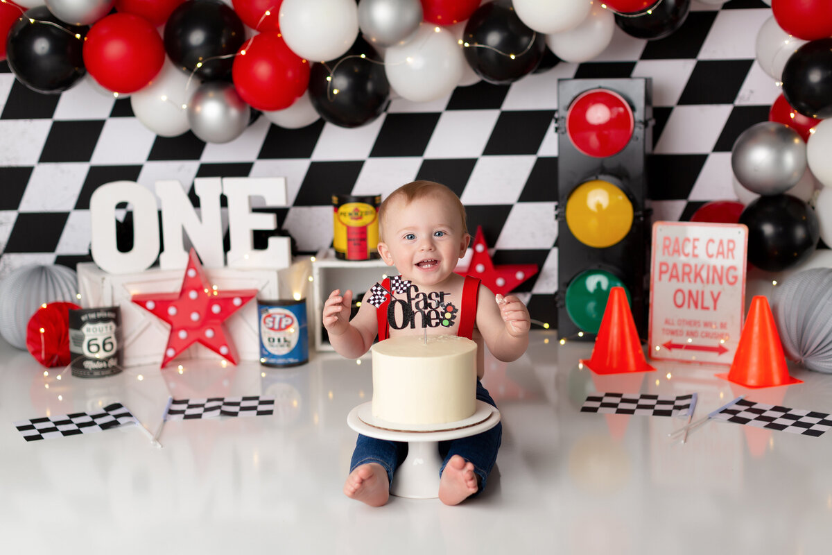 Racecar Nascar themed cake smash at West Palm Beach, FL newborn photography studio.  Baby boy sitting behind untouched white cake smiling at the camera. In the background, there is a black and white checkered backdrop, black, white, and red balloons, a stop light, and race flags.