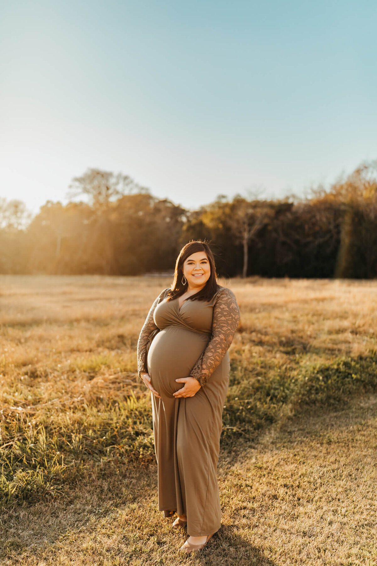 Expectant mother holding her belly in an olive lace dress in a mowed field.
