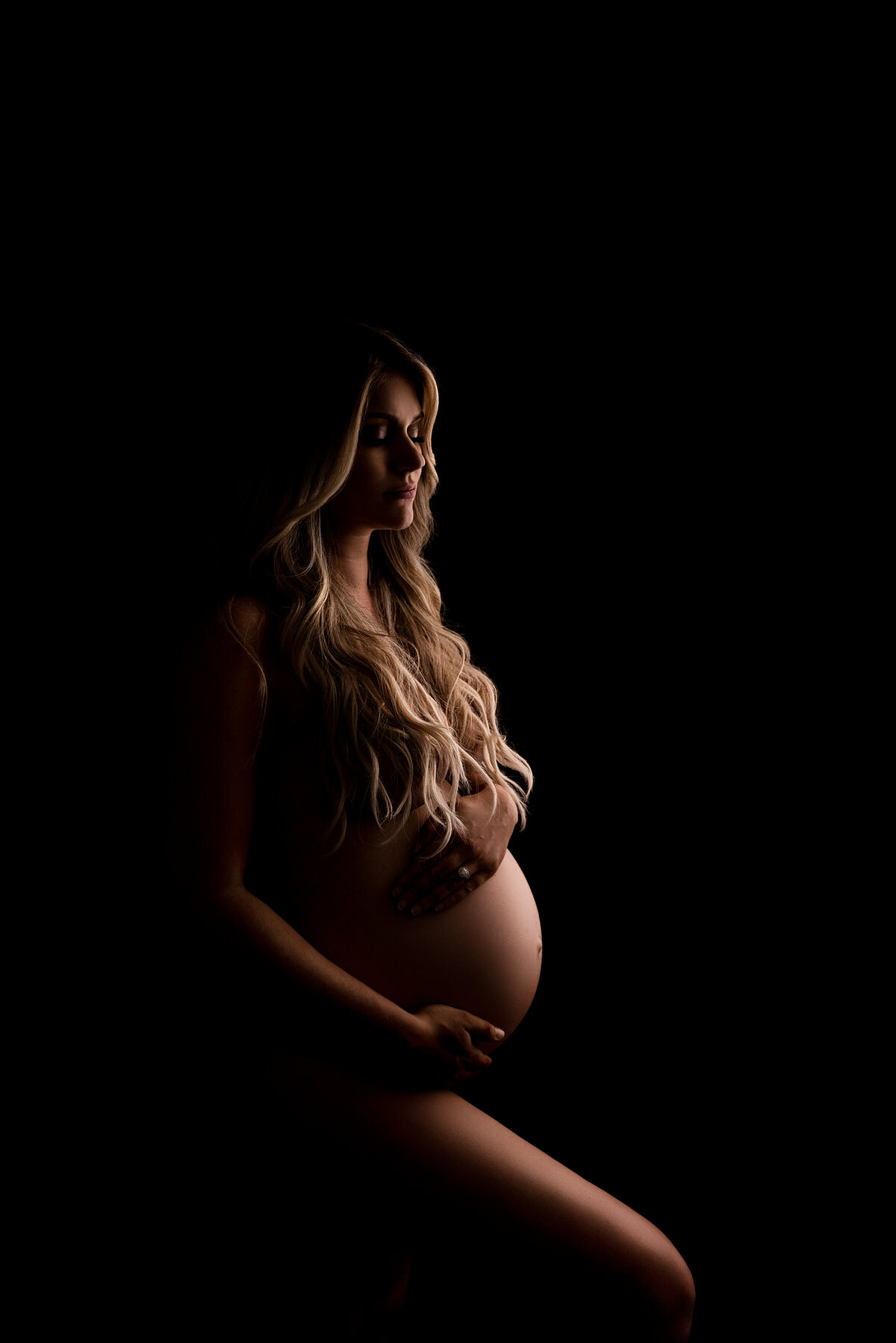 Philadelphia Main Line's best maternity photographer Katie Marshall captures expectant mom for a fine art maternity photoshoot. Bare photo of an expectant mom with dramatic lighting - only parts of mom are highlighted, the rest is shadowed black. Mom's leg is bent in front of her, her hands are gently resting on her bump.