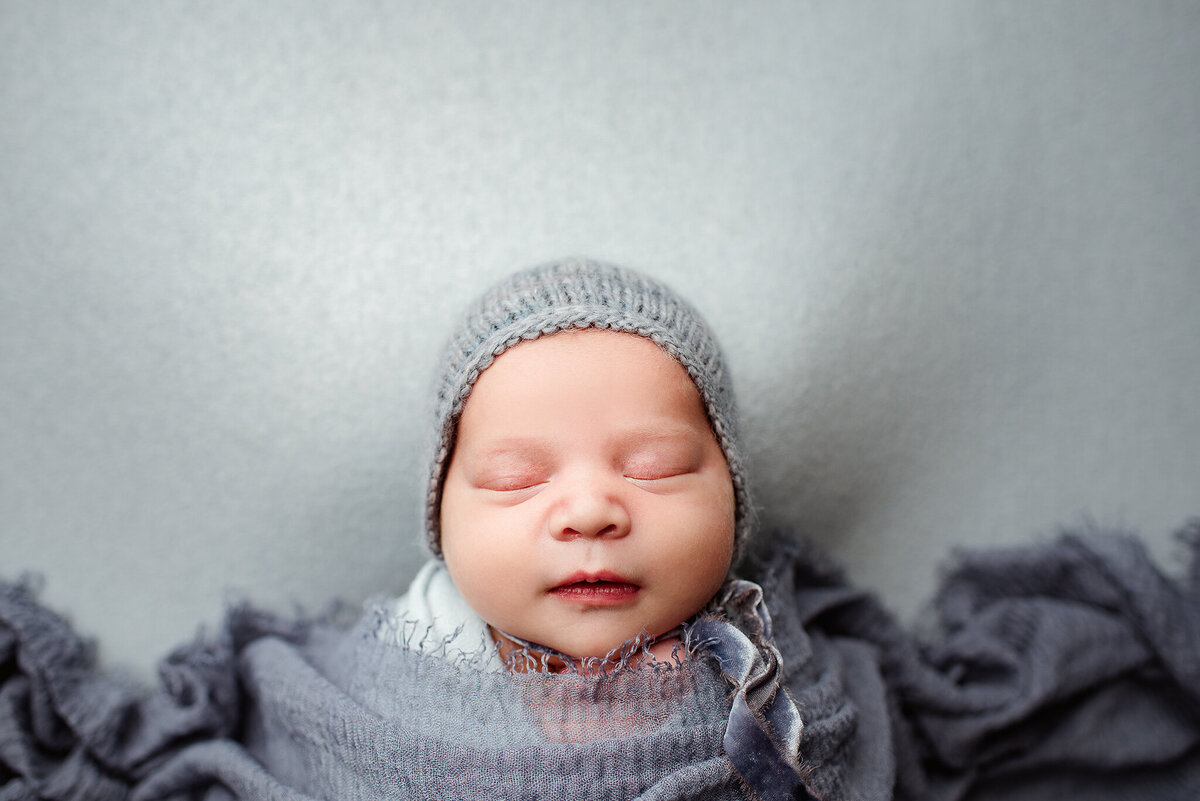 In home studio newborn portrait of baby boy in blue bonnet and wrap in Jacksonville, FL.