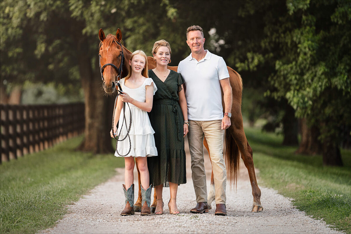 young blonde equestrian wearing white dress and cowboy boots standing next to her mother in a green dress, her father in a white shirt and khaki pants, with their chestnut thoroughbred horse behind them. They are standing on a gravel path that is lined with green grass and trees behind them.