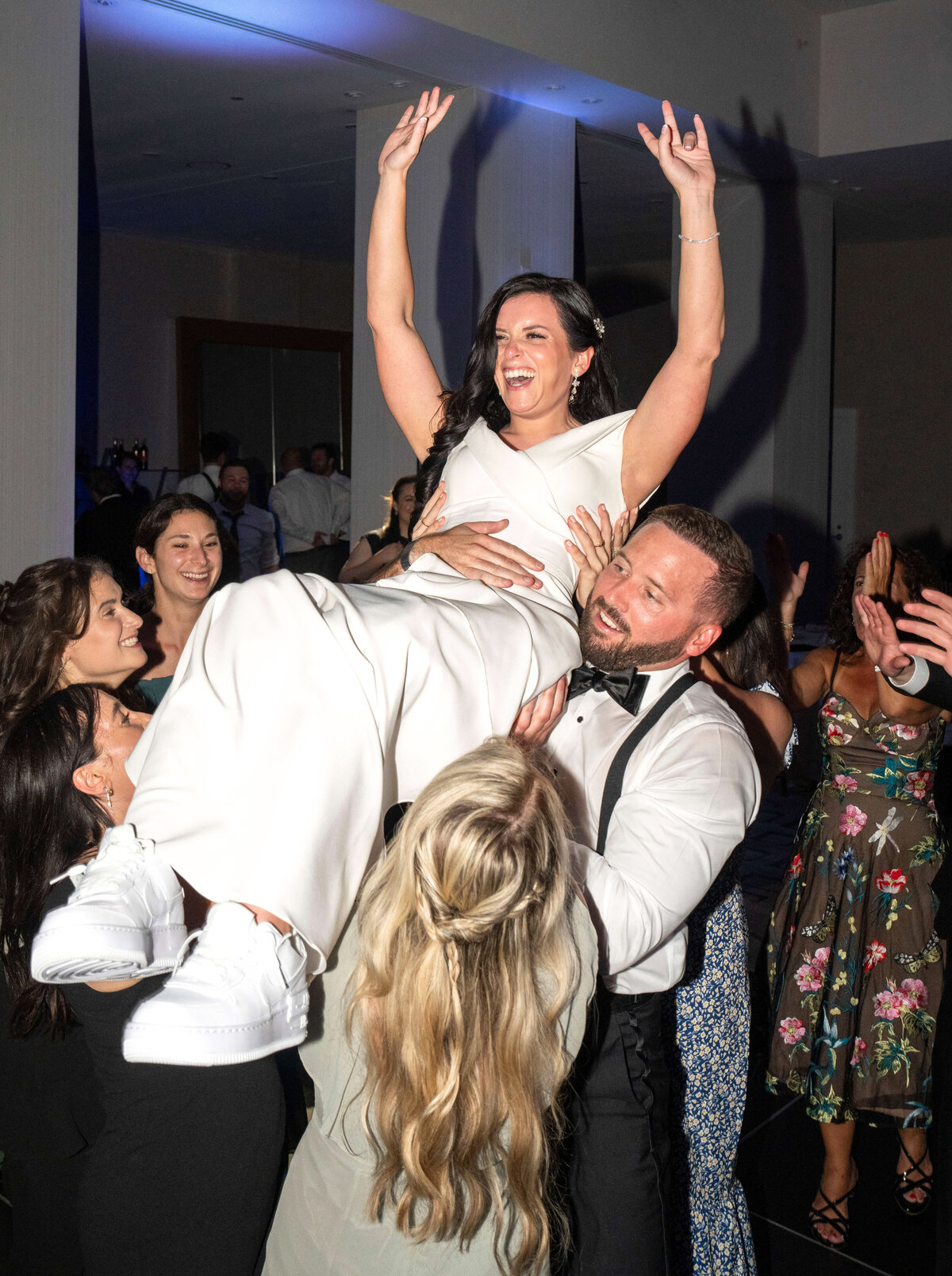 A joyful bride in a white dress and sneakers is lifted into the air by guests at a lively celebration. The crowd around her is cheering and clapping, creating a festive atmosphere.