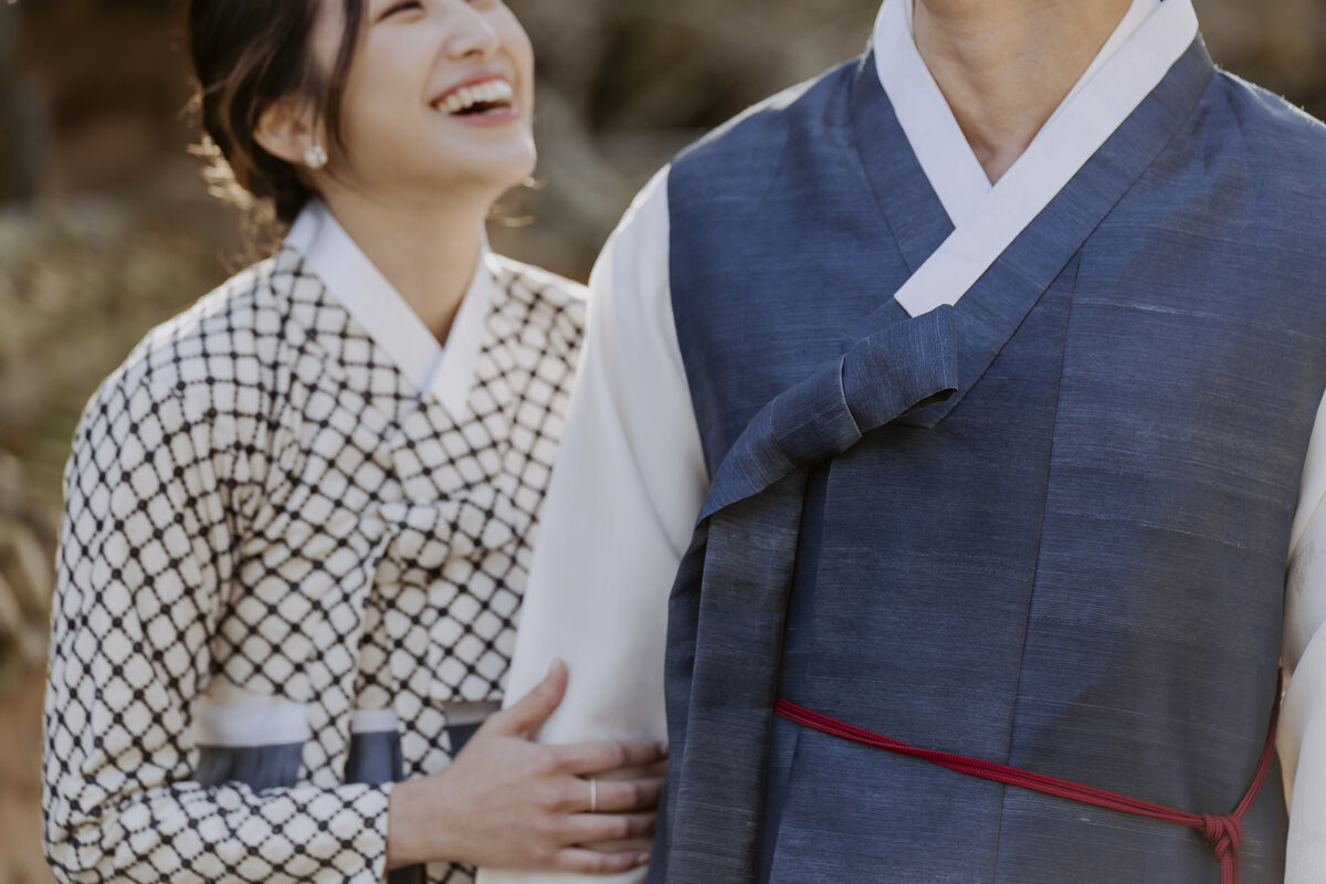 the bride is laughing at the groom while wearing a checkered hanbok with blue ribbon while the groom wears a blue and white with red ribbon