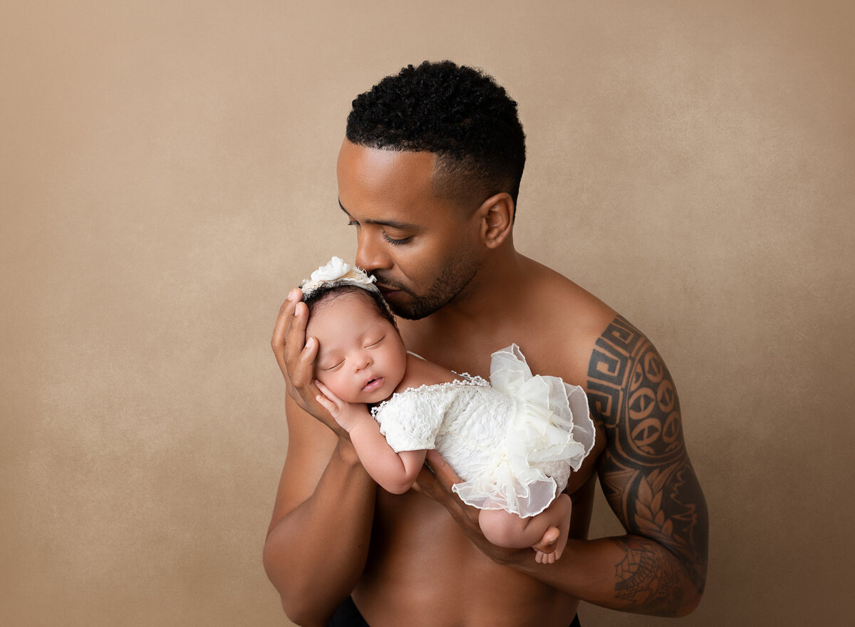 Shirtless new dad is holding his new baby girl in his arms. Baby is laying on her belly on dad's forearm and the other hand is under her head. Dad is smelling baby's hair.
