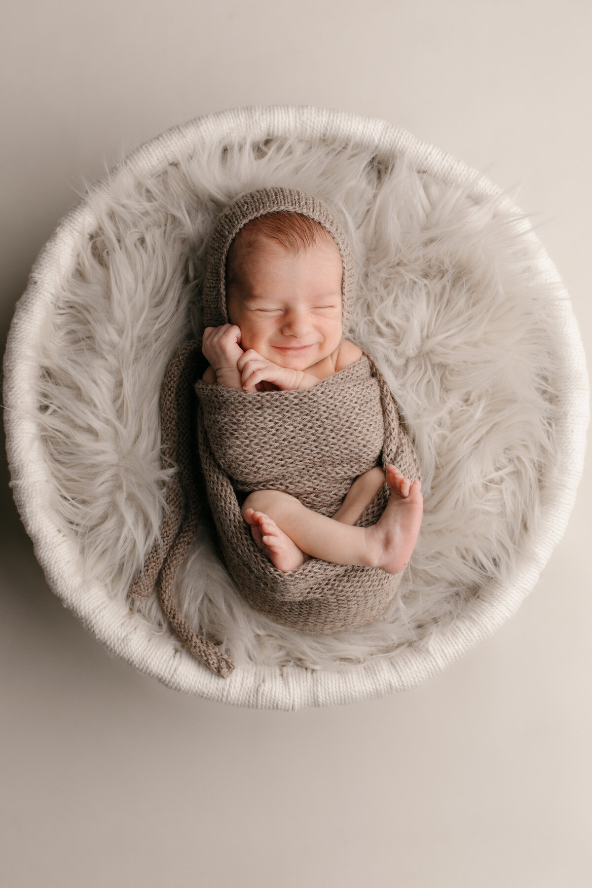 Portrait of a newborn baby swaddled sweetly in a tan wrap, sleeping and smiling while set on a fur lined basket in Los Osos, Ca photography studio