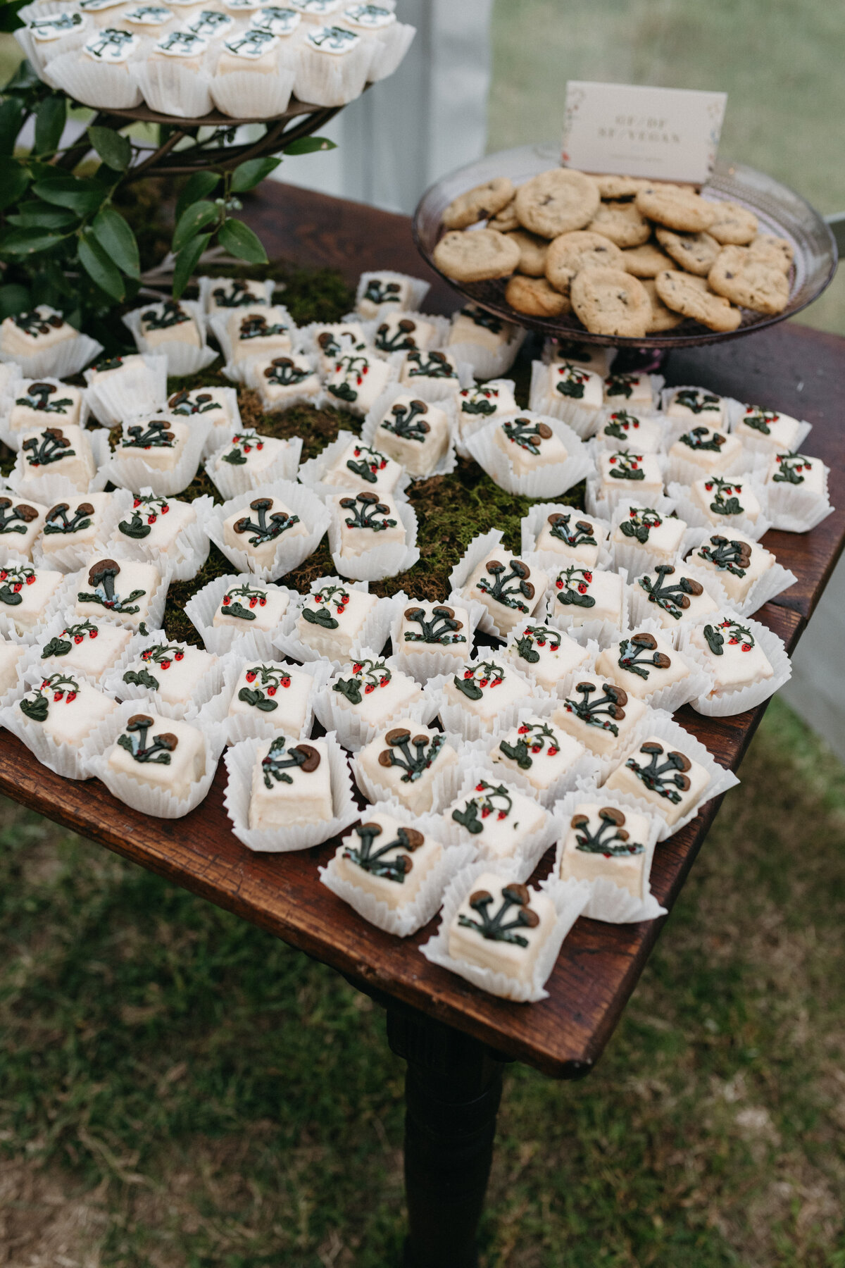 Whimsical dessert table featuring hand-decorated mushroom petit fours and cookies displayed on moss for an enchanted woodland-inspired wedding at The Nest in Northwest Arkansas. Styled details complement the garden-style floral aesthetic by Fleurish Floral Studio.