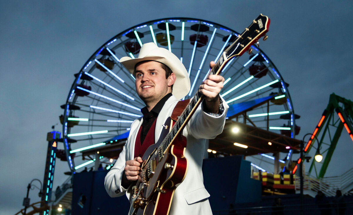 Male musician portrait Ben Klick wearing white suit white cowboy hat while holding outstretched red  electric guitar with ferris wheel behind