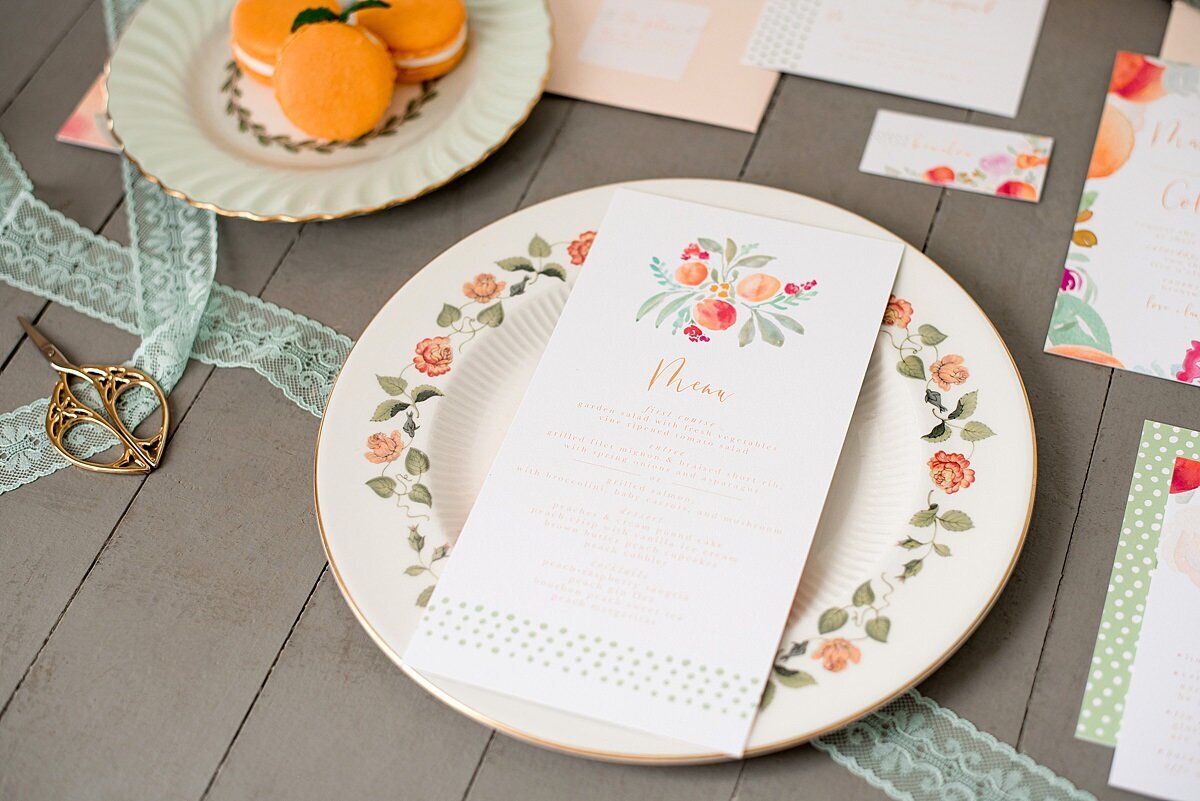 Mismatched vintage china plates with peach macarons and peach themed wedding stationery set. Wedding menu card decorated with peaches, pink flowers and green leaves.