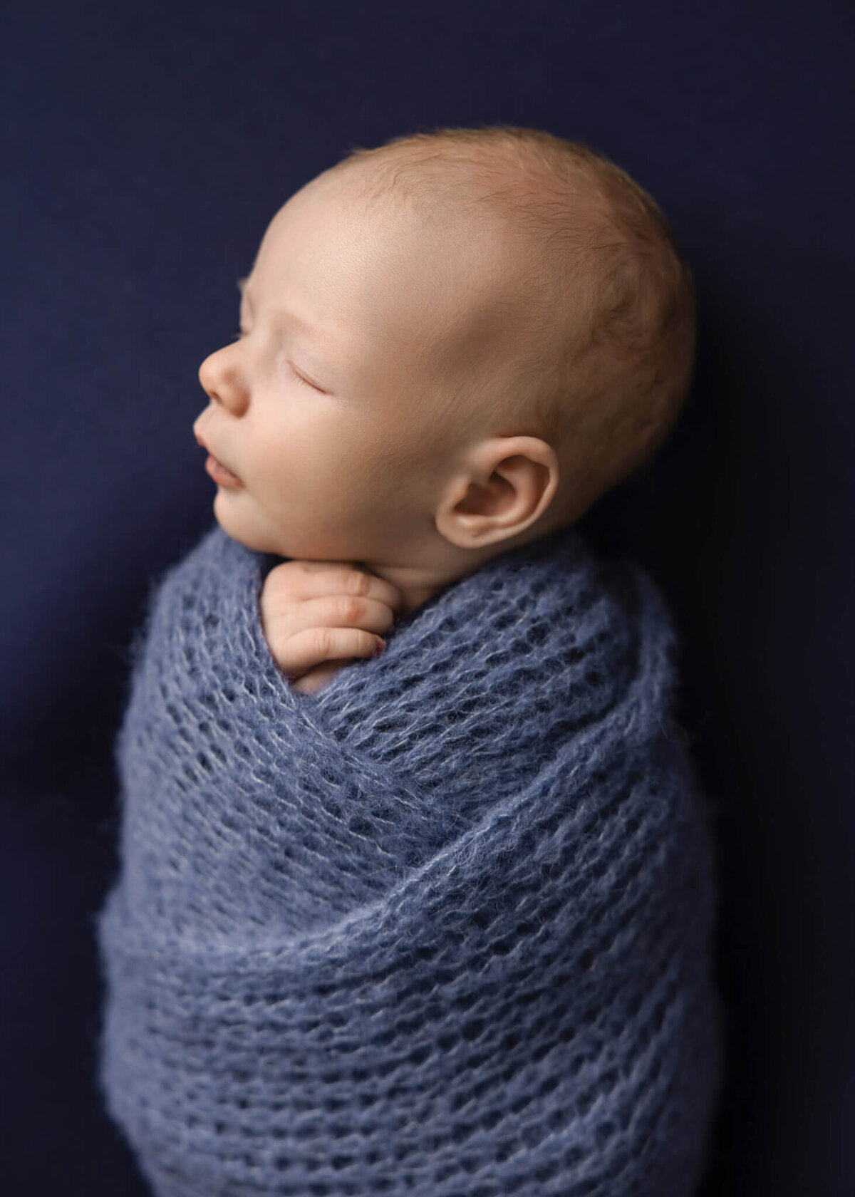 close up of newborn baby wrapped in blue kntted wrap asleep on blue fabric
