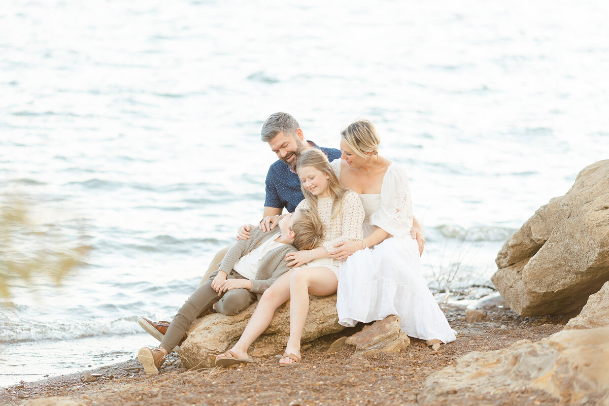A beautiful lake side family photo of the family of 4 sitting on a rock as they interact with each other as their Fort Worth family photographer captures the moment.