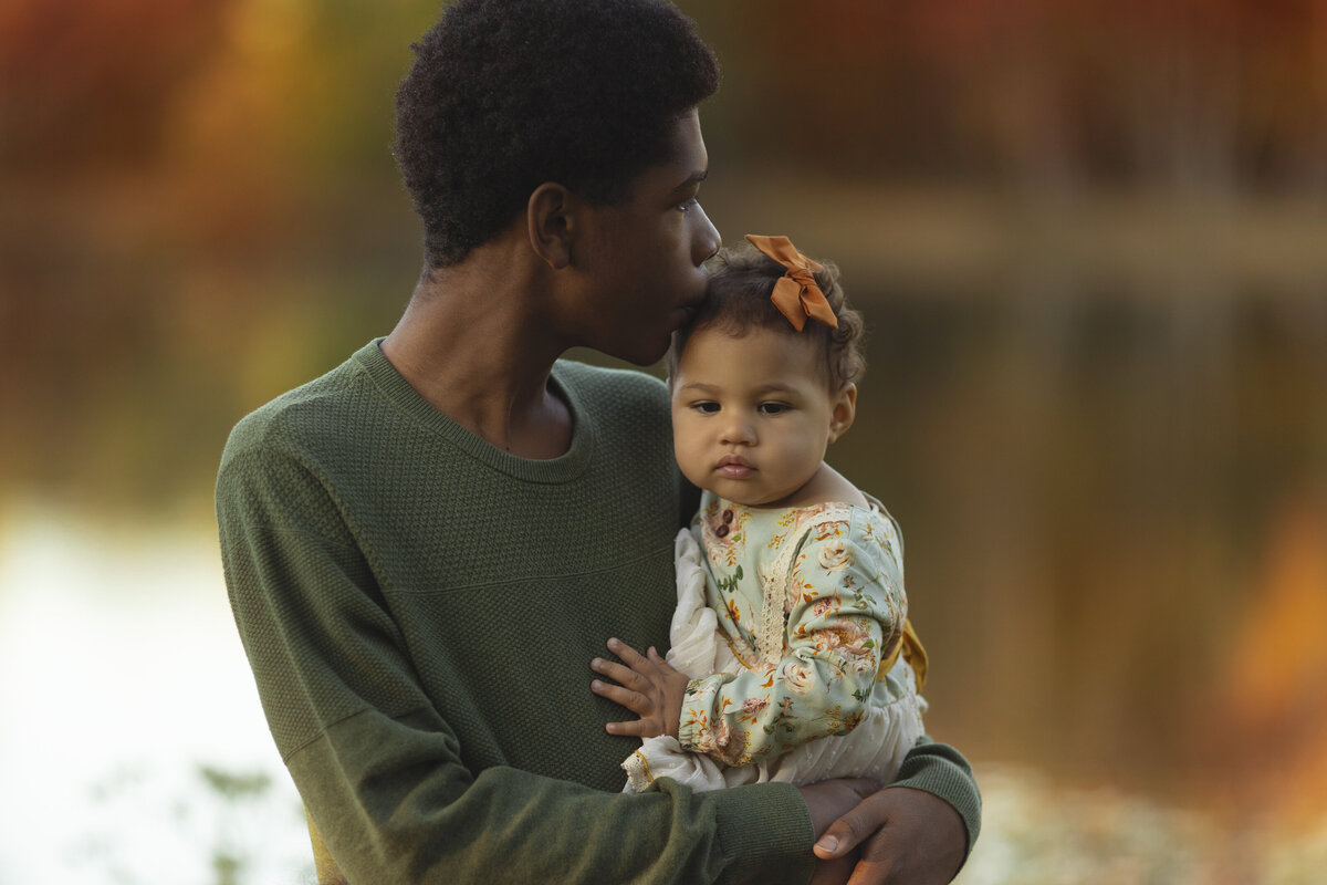 An image from a New Jersey Family Photographer of a teenage boy kissing the head of his toddler baby sister in his arms while standing by a lake