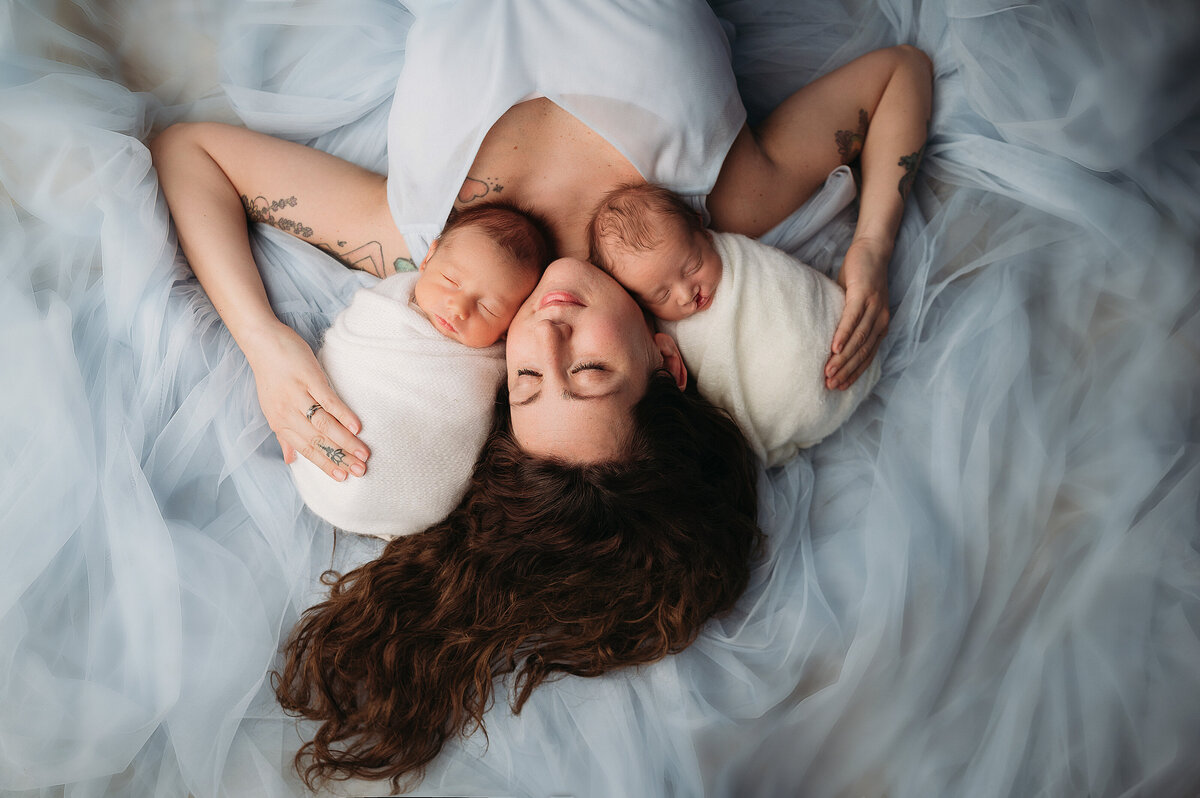 mom with brown hair laying on grown in blue gown holding her newborn baby boys wrapped in white swaddles son with cleft lip and palette