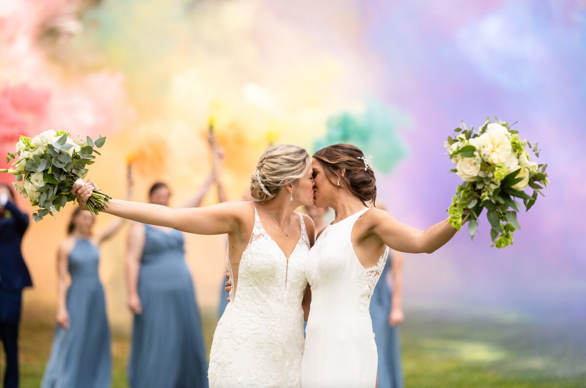 two brides kissing with their bridal party behind them holding rainbow smoke bombs on the grounds of the legacy at woodcrest