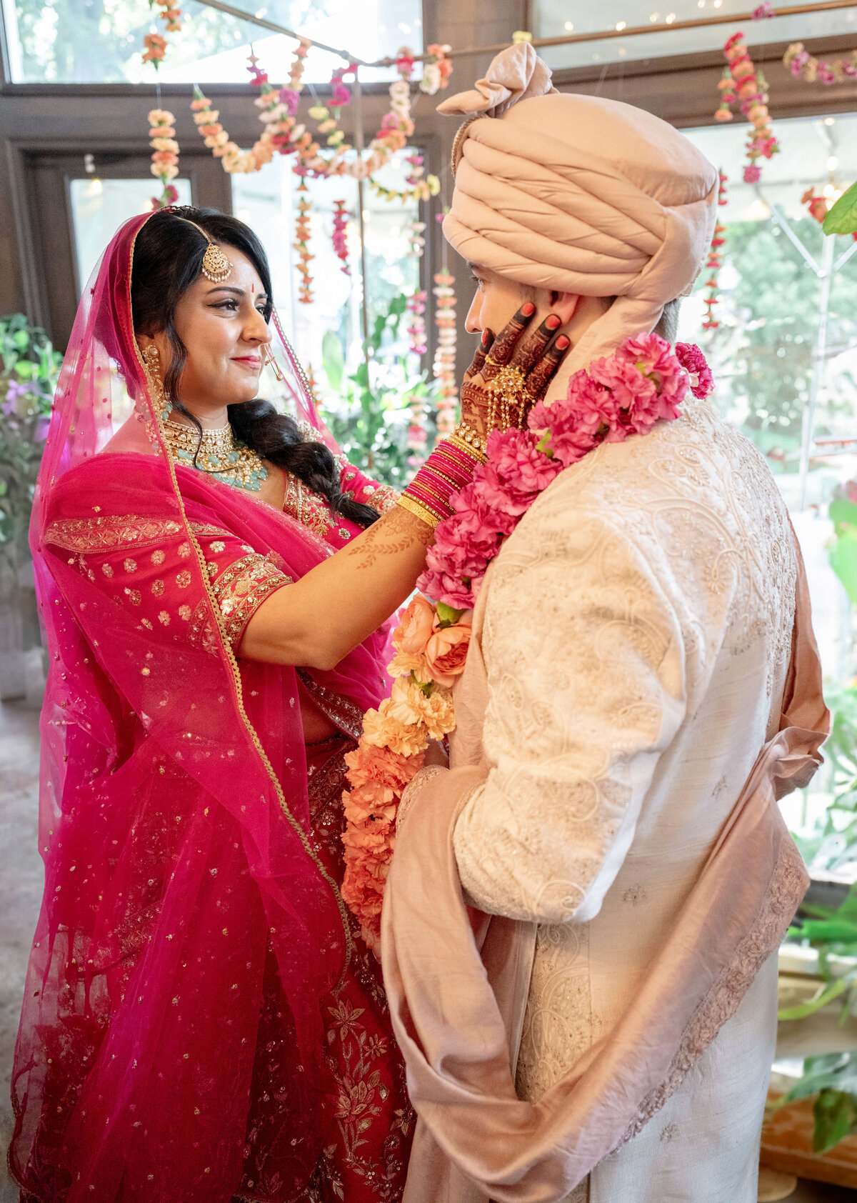 A bride in a vibrant pink traditional outfit tenderly touches the grooms face, who is dressed in an ivory sherwani and turban. Both wear pink flower garlands. The background is adorned with colorful floral decorations.