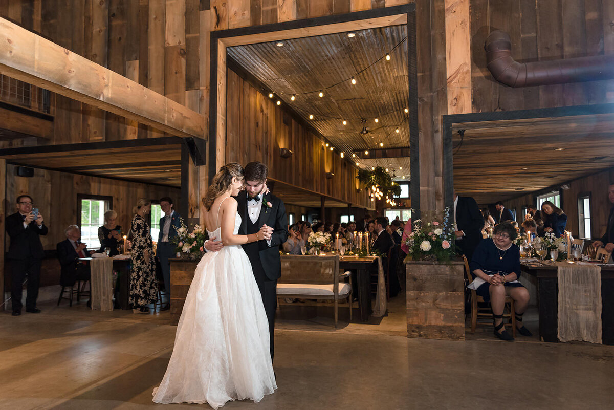 A bride and groom sharing their first dance in a rustic barn with guests watching from their tables.