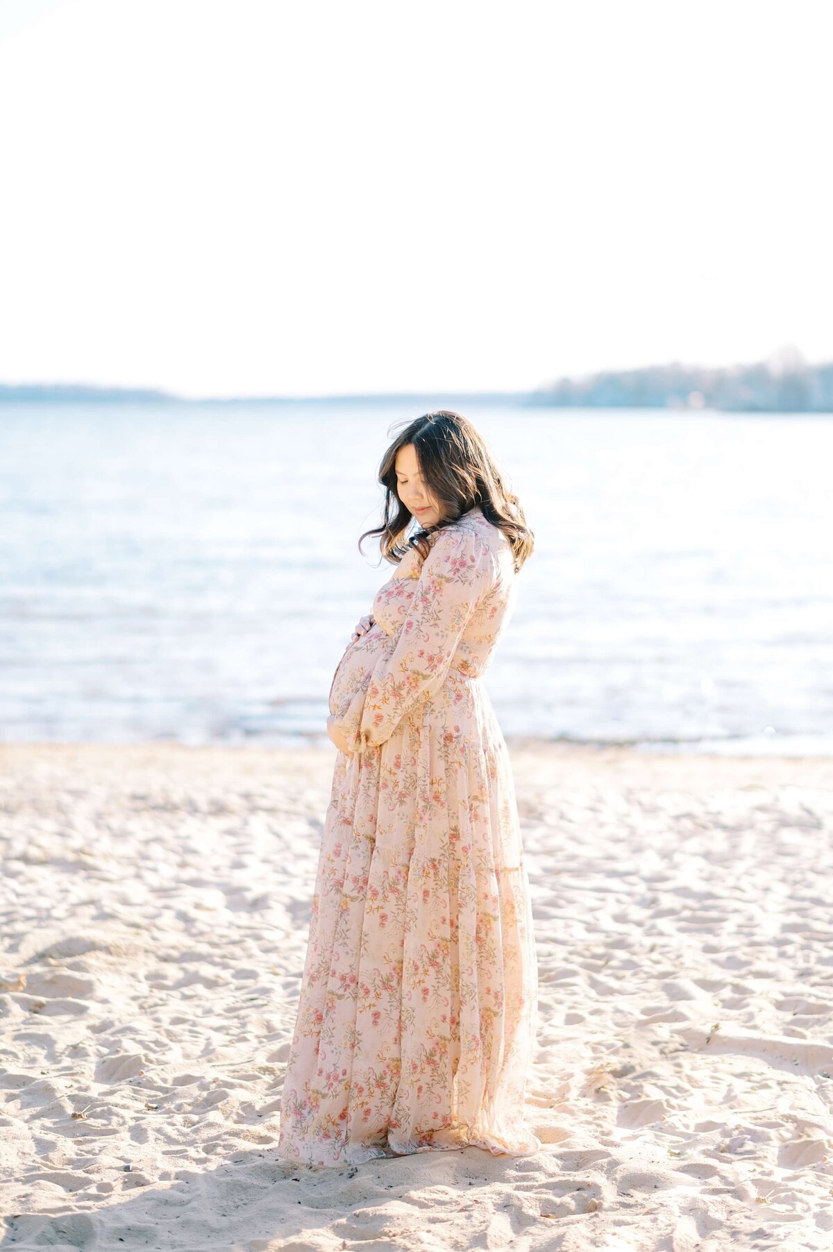 Charlotte Maternity Photographer, Melissa Mayrie Photography captures an expecting mama on a windy beach while cradling her belly in a pink floral dress.