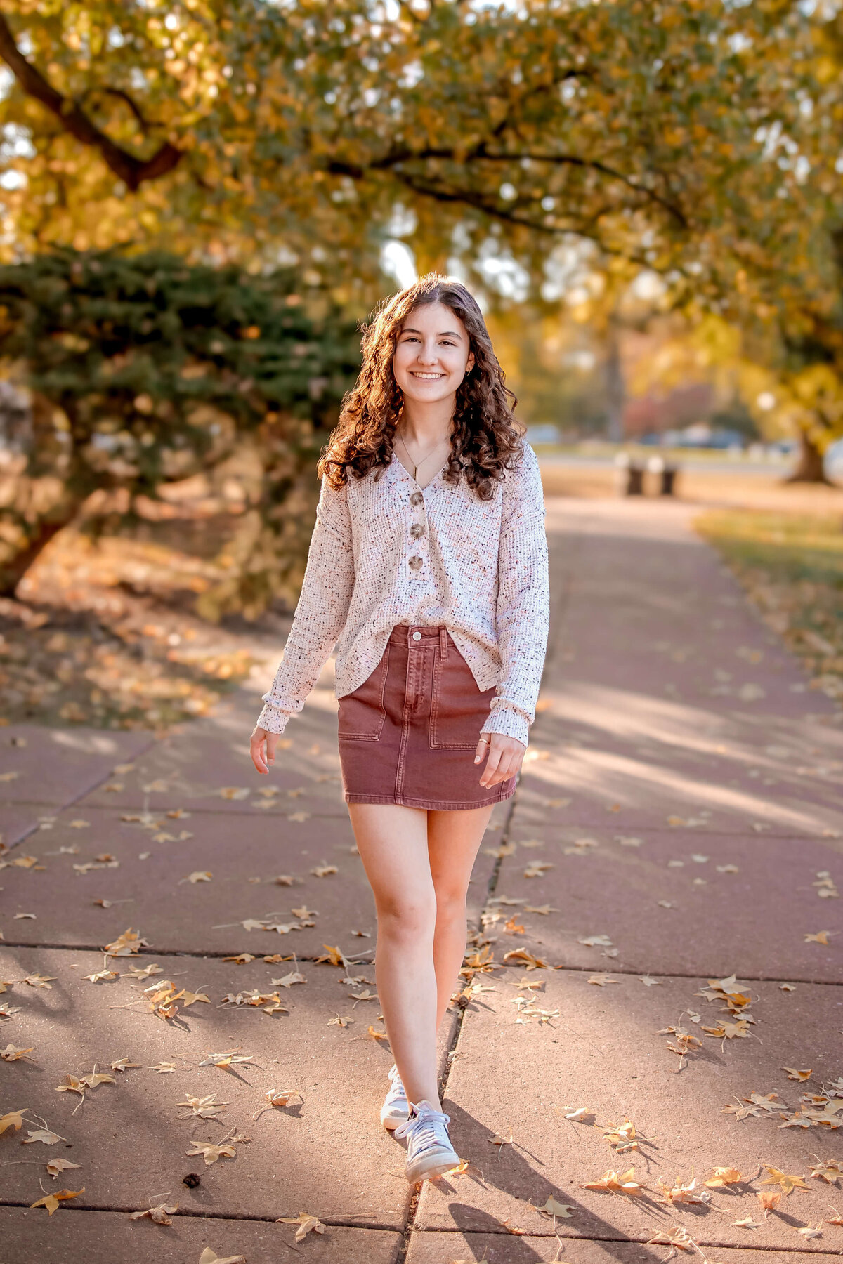 A gorgeous young girl is walking towards the camera in a park filled with fall leaves and beautiful colors.