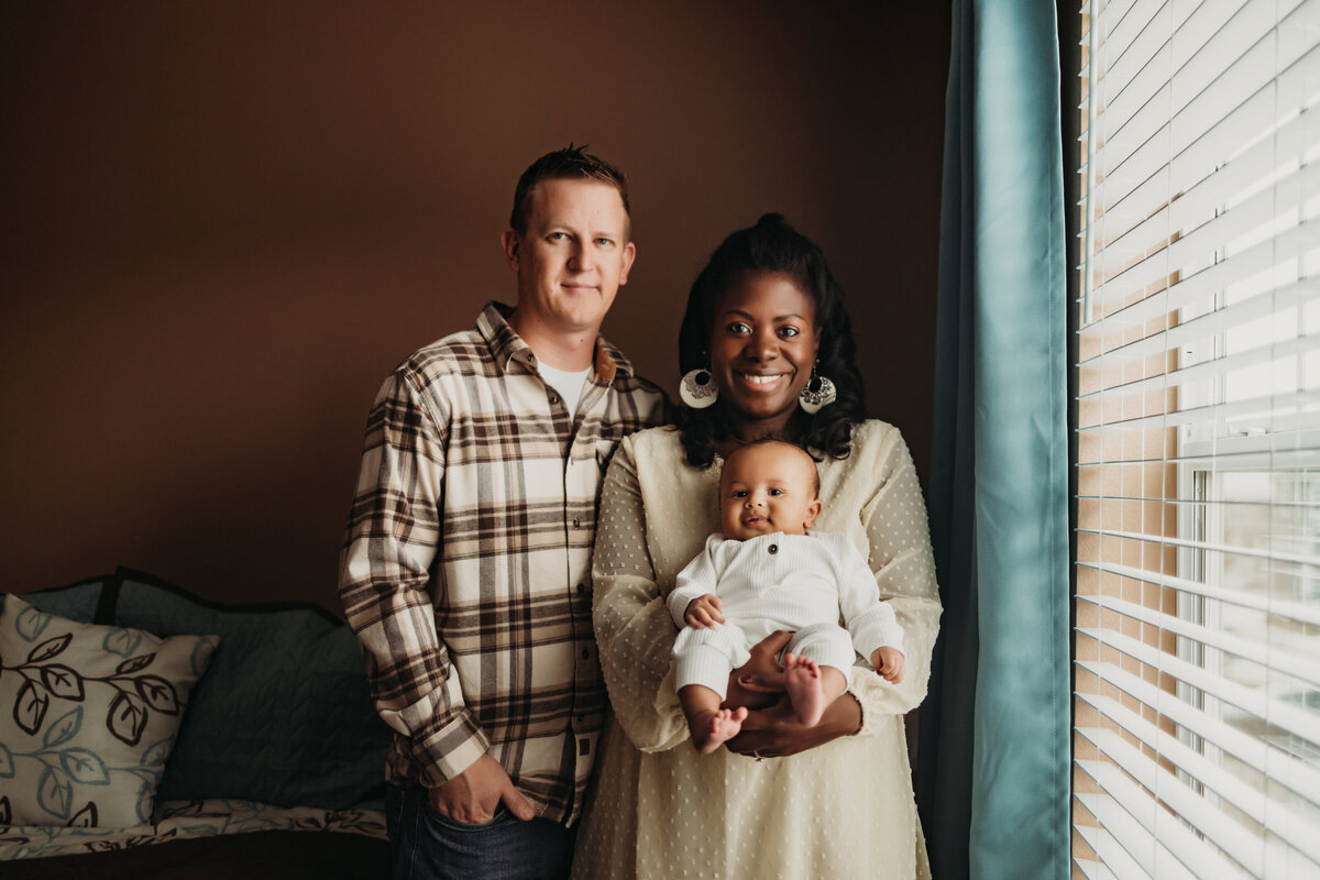 famly of three holds newborn baby boy by window in  master bedroom