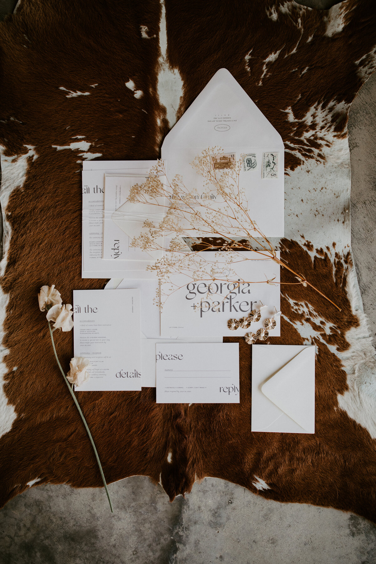 Various white wedding stationery with black font atop cow hide with dried stems and a flower.