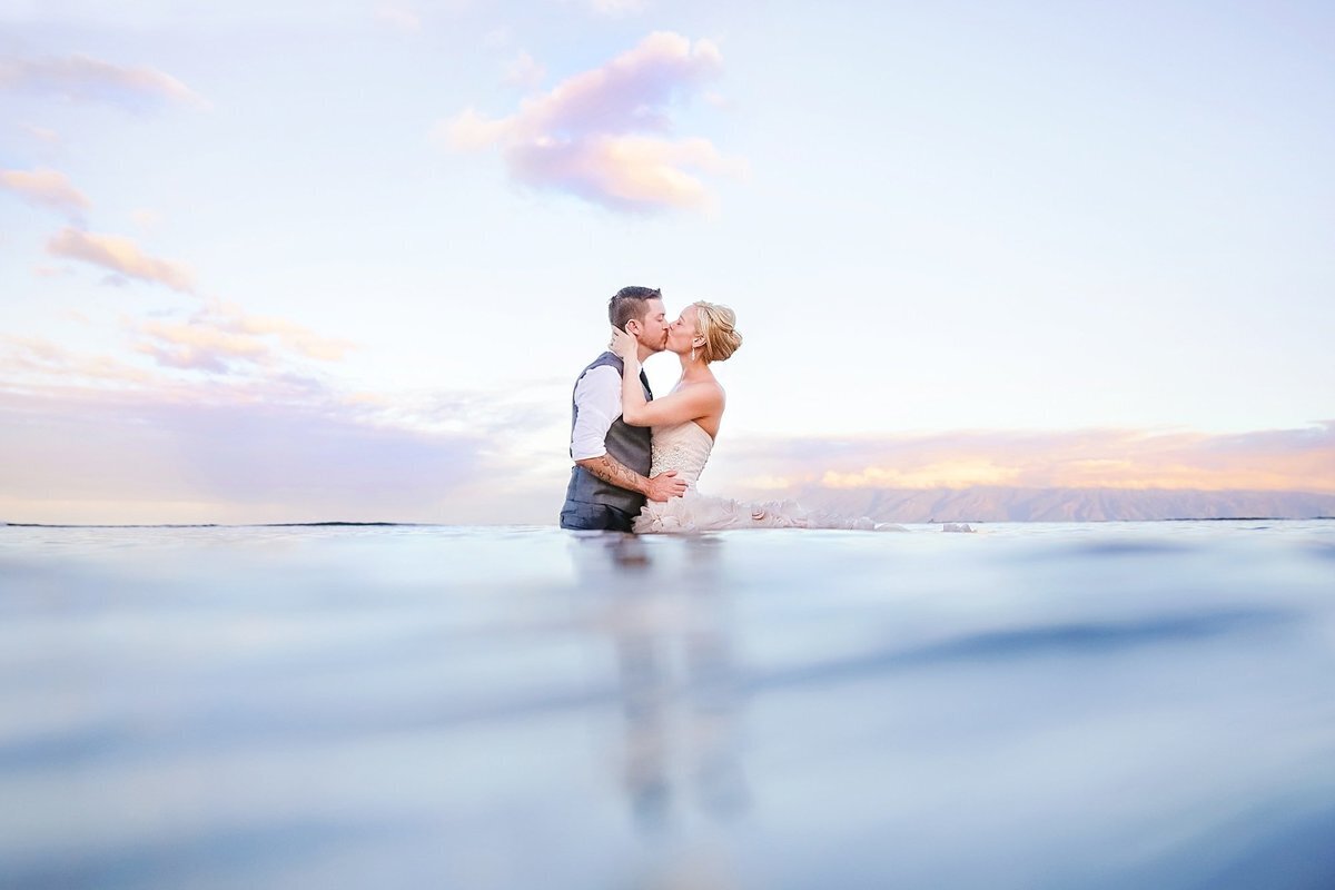 Woman with her hair in a bun wearing a pink fluffy wedding dress kisses her new husband in the ocean while being photographed by Love + Water