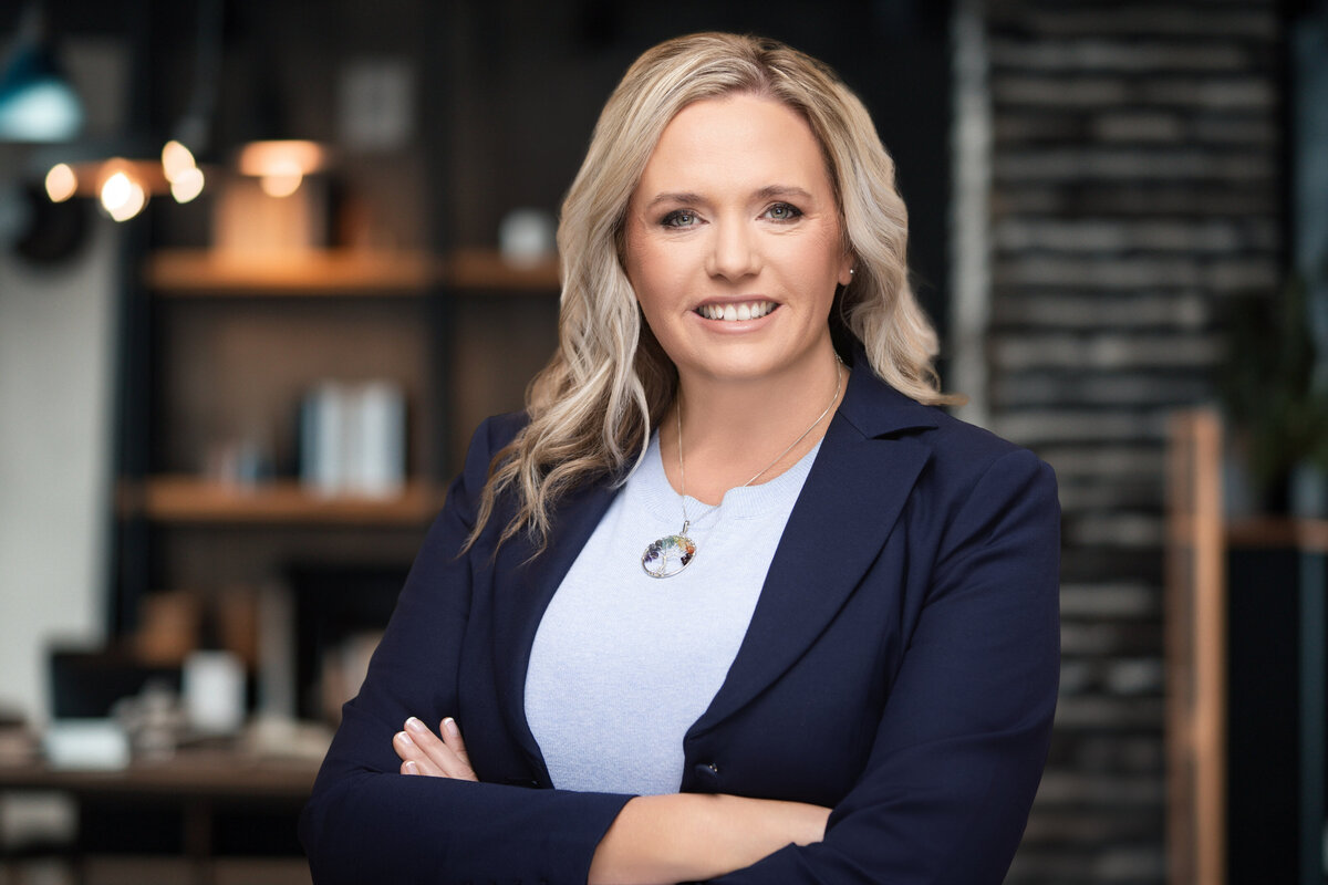 Authentic and approachable professional headshot of a blonde woman with soft curls, wearing a tailored navy blazer over a light blue top, photographed in a modern office space with warm, ambient lighting. Captured at Janel Lee Photography, a Cincinnati headshot studio known for elevated branding portraits for executives, HR leaders, and corporate professionals. This image is ideal for speaking engagements, LinkedIn banners, press features, and professional networking platforms across the Midwest.
