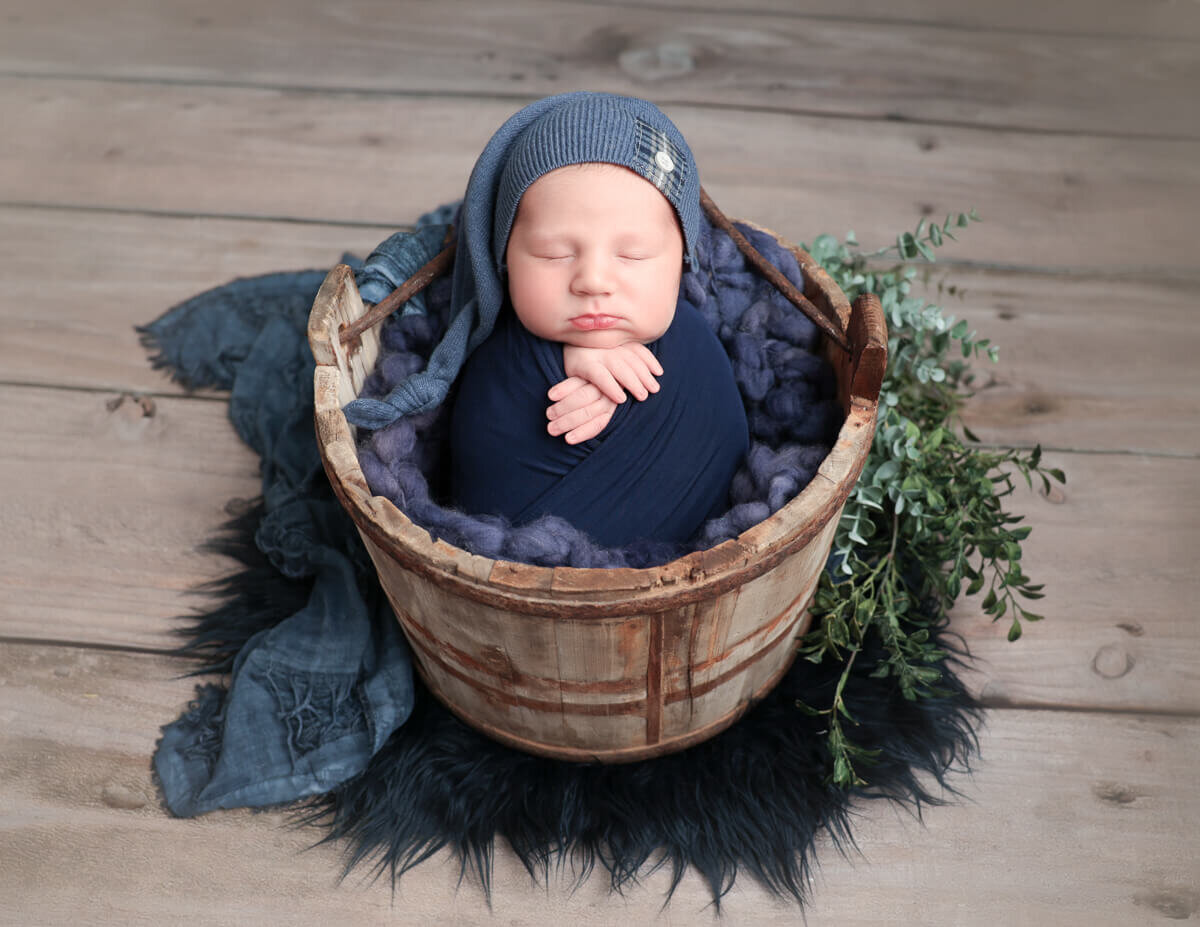 A peaceful newborn boy wrapped in a navy blanket, nestled in a woven basket surrounded by soft greenery, captured in a photography studio in Rochester, NY.