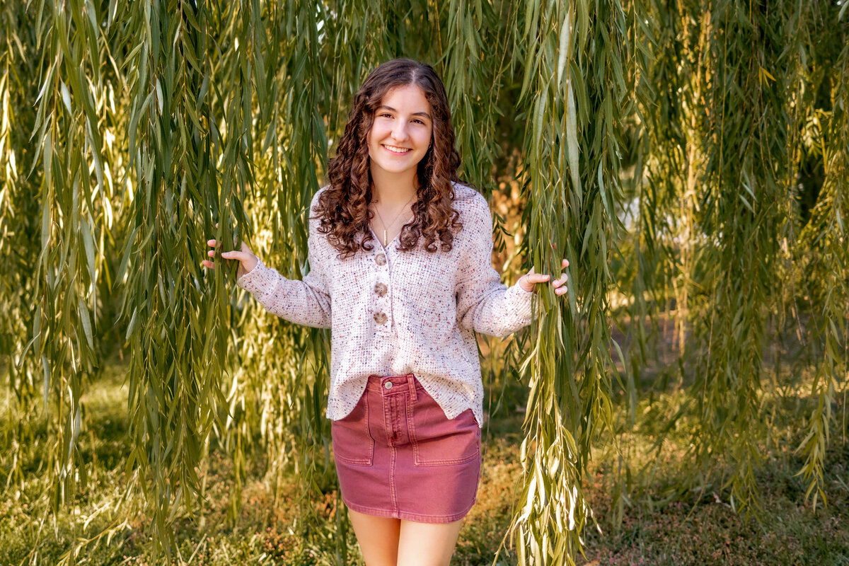 A beautiful girl is stading in a willow tree and peeking out from the long leaf vines, smiling nice for her senior photos with Foppiano Photography.