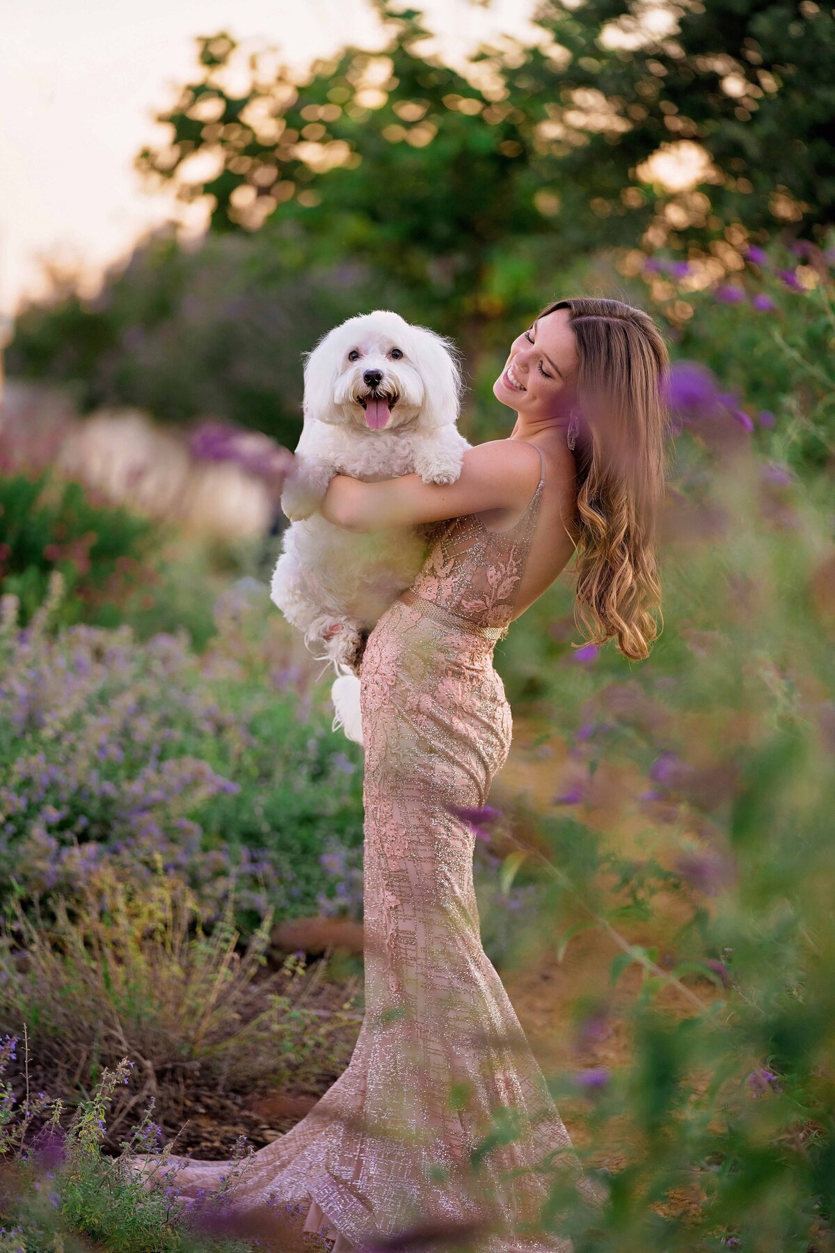 Young woman wearing a pink prom dress poses in a lavender field holding her white fluffy doodle dog.