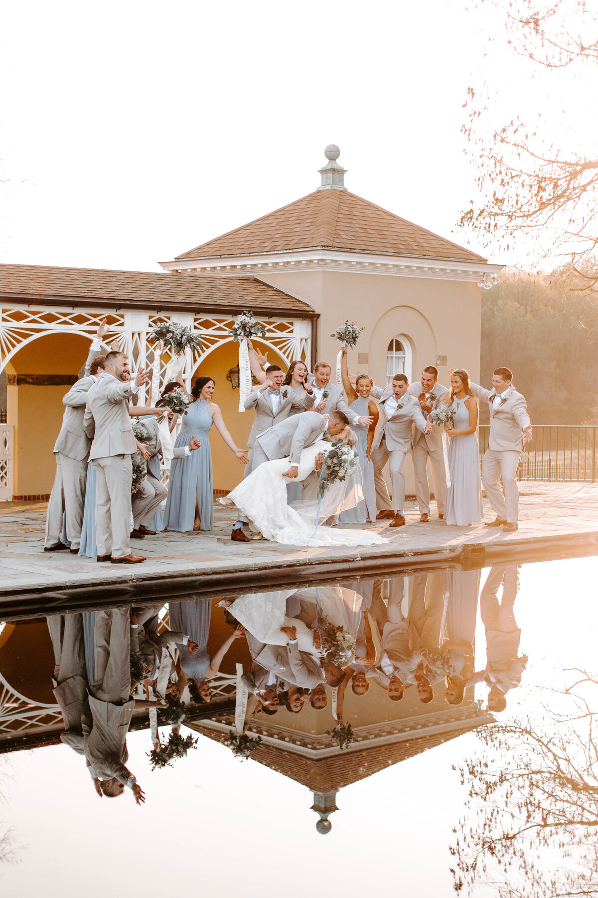 A bride and groom are kissing as the bride is being dipped, they are surrounded by their wedding party who are all cheering. The group stands behind a pool that perfectly reflects the image