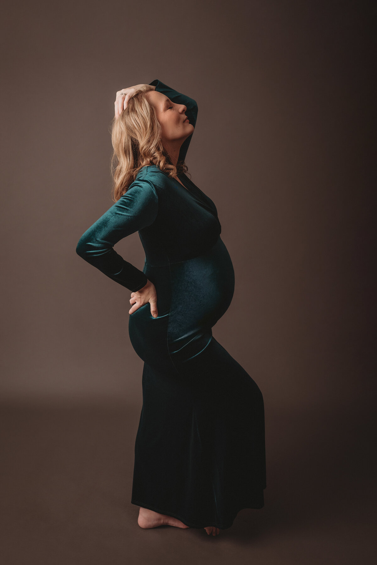 pregnancy picture of woman 37 weeks pregnant wearing green velvet dress  posing with hand on back and one leg popped up on dark gray backdrop