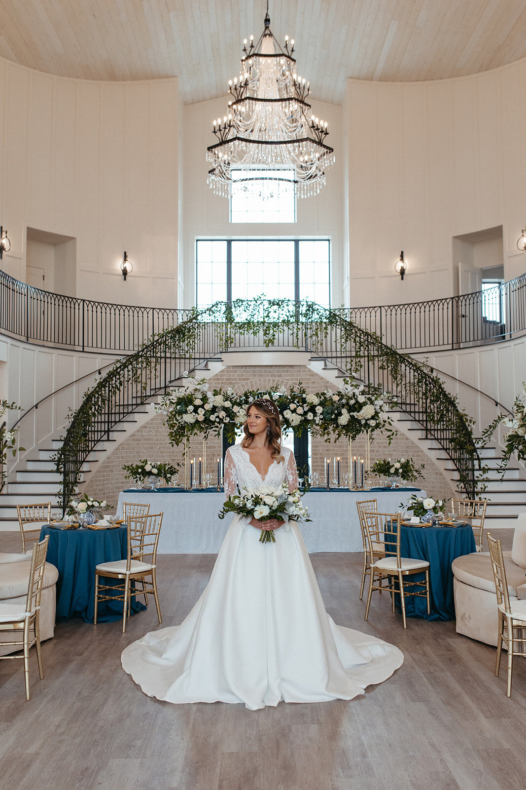 A bride in a white wedding gown holds a bouquet below chandelier in front of a two-sided staircase wrapped in garland.